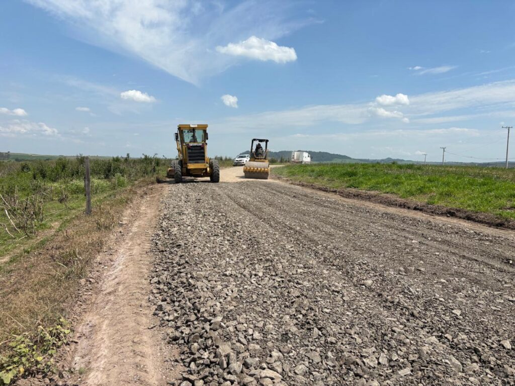 Estrada na zona rural recebendo cascalho, assentado por um trataor amarelo e um rolo compressor com detalhe amarelo.