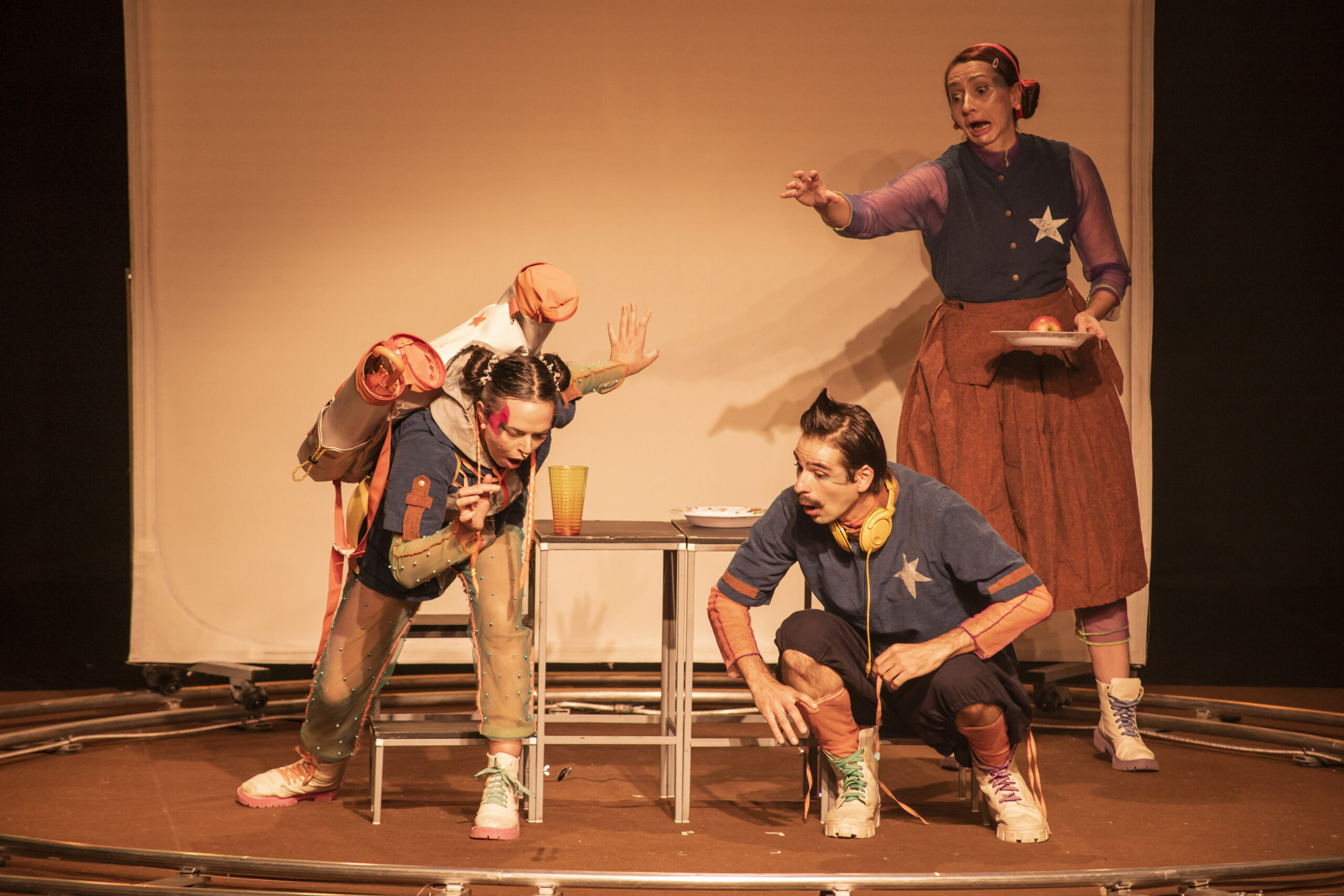 Três atores atuando, caracterizados, em um cenário de luz amarela