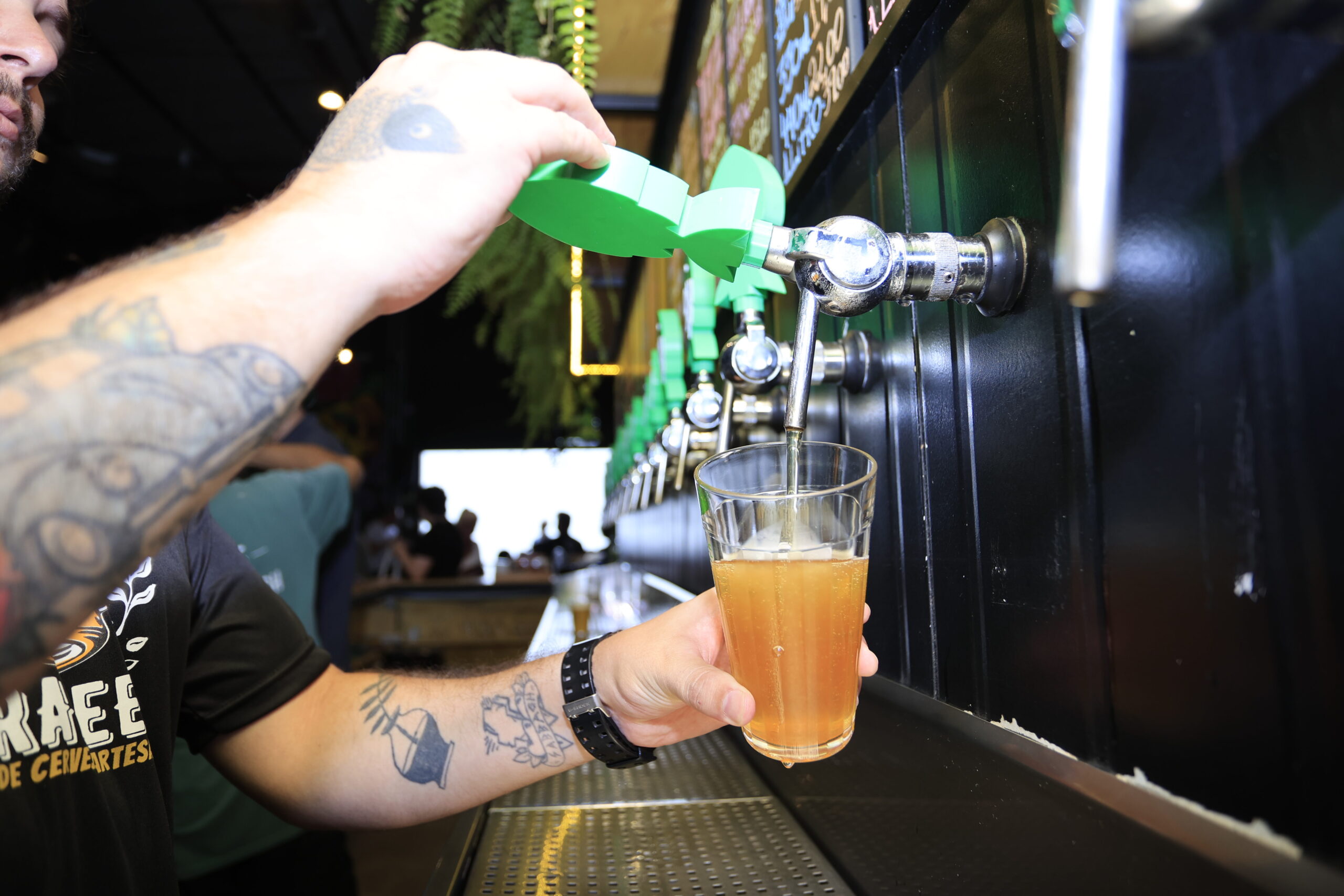 A imagem mostra um garçom com tatuagens nos braços despejando cerveja de um sistema de torneiras em um copo. A mão do garçom segura um dispositivo verde plástico que parece direcionar o fluxo da cerveja para o copo. O copo é de vidro transparente e está quase cheio de uma cerveja âmbar. A torneira de onde a cerveja está sendo servida é metálica e brilhante, e faz parte de uma fileira de outras torneiras semelhantes. O fundo é escuro e um pouco desfocado, com alguns clientes visíveis ao fundo do bar. O garçom está usando uma camiseta escura com um logotipo pouco visível. A imagem é bem iluminada, com foco principal no ato de servir a cerveja. A cena sugere um ambiente de bar ou cervejaria artesanal, descontraído e informal.