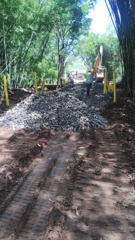 Ponte na zona rural recebendo pedregulho em sua base, tendo ao fundo dois funcionários e uma máquina motoniveladora.