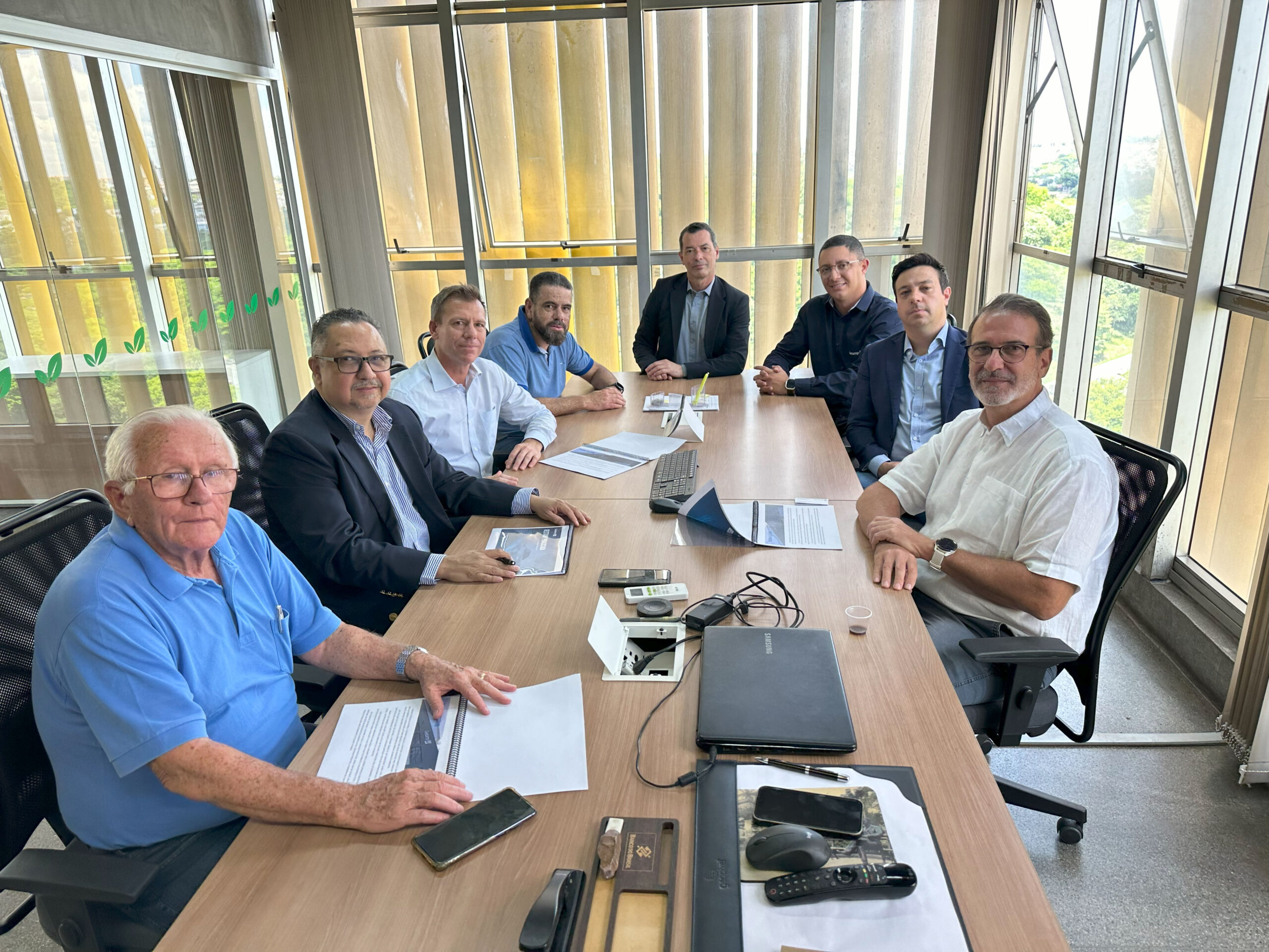 A imagem mostra uma reunião de oito homens em uma sala de conferências moderna e bem iluminada. Eles estão sentados em torno de uma grande mesa de madeira retangular. A sala tem grandes janelas de vidro que oferecem uma vista panorâmica da paisagem externa. A mesa está organizada com pastas, laptops, telefones celulares e outros objetos relacionados a negócios. Os homens parecem estar envolvidos em uma discussão ou apresentação de negócios, com documentos espalhados na mesa. Eles estão vestidos de forma profissional, com camisas sociais e blazers. A atmosfera parece formal, mas profissional e colaborativa. A iluminação é brilhante e natural, vindo das grandes janelas. O espaço é limpo e organizado, refletindo um ambiente de trabalho moderno e eficiente. O piso é claro e o teto é alto, contribuindo para a sensação de espaço e amplitude. No geral, a imagem sugere uma reunião de negócios importante em andamento.