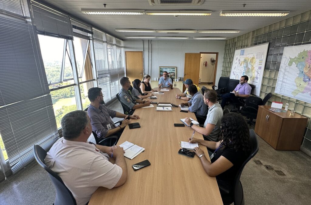 Imagem de uma sala com uma mesa grande e várias pessoas sentadas à mesa. Ao fundo, o prefeito Luciano Almeida com camisa azul