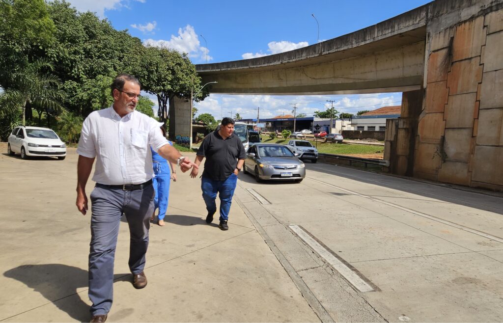 Em primeiro plano, o prefeito usando camisa branca e calça jeans, acompanhado do engenheiro que usa camiseta preta e calça jeans; do lado direito da imagem, um trecho da avenida já pavimentada em concreto e sinalizada