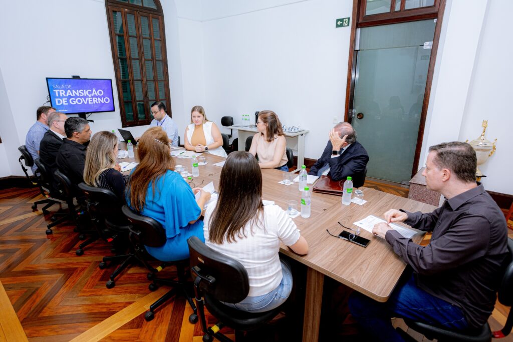 A imagem mostra uma sala de reuniões com uma longa mesa de madeira onde várias pessoas estão sentadas. A sala é bem iluminada e parece formal. Na parede, há uma tela mostrando a frase "Sala de Transição de Governo". A mesa está repleta de garrafas de água e copos, indicando que uma reunião está em andamento. As pessoas estão sentadas em cadeiras giratórias com encosto alto. Há mais homens do que mulheres na reunião, e a maioria deles está vestindo roupas formais. Uma mulher está sentada no canto da mesa com uma expressão de aparente preocupação e um homem está sentado ao lado dela de cabeça baixa. O chão é de madeira escura com um padrão em espinha de peixe. A atmosfera parece séria e profissional, sugerindo uma reunião de trabalho importante, possivelmente relacionada à transição de governo.