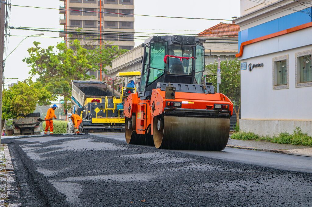 Máquina com detalhes laranja trabalha na pavimentação de avenida.
