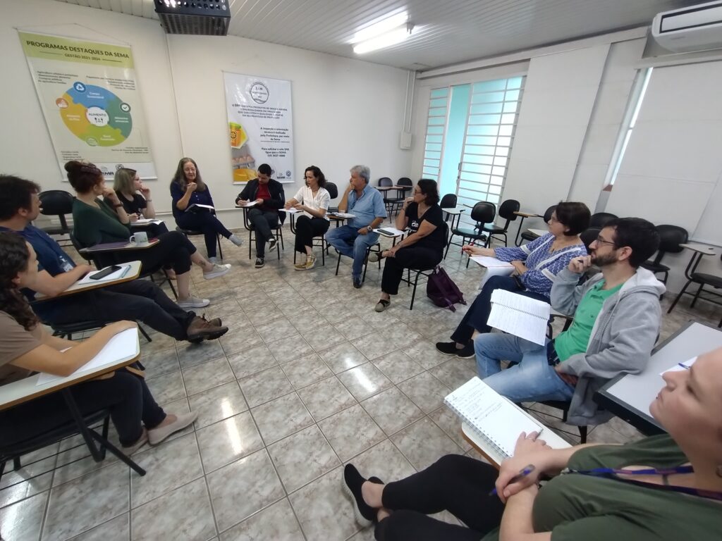 A imagem mostra um grupo de pessoas sentadas em cadeiras em círculo numa sala de aula ou sala de reuniões. Elas parecem estar participando de uma reunião ou workshop. Há várias carteiras com cadernos e canetas sobre elas, sugerindo que estão tomando notas. A sala tem paredes claras, um piso de cerâmica e algumas janelas que deixam entrar luz natural. Na parede, há dois cartazes, um deles parece descrever programas e o outro, uma instituição ou organização. As pessoas parecem estar envolvidas em uma conversa ou discussão. A atmosfera é calma e profissional.