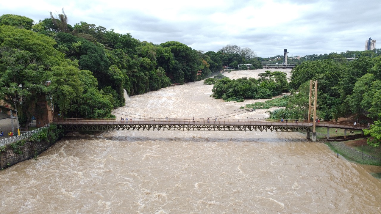 A imagem mostra uma ponte de madeira suspensa sobre um rio caudaloso e turvo. A água está marrom e agitada, indicando um fluxo forte. A ponte tem um deque de madeira e cabos de suporte visíveis, com várias pessoas caminhando sobre ela. As margens do rio são densamente arborizadas, com árvores verdes exuberantes que se estendem até o fundo da imagem. Existe uma cachoeira pequena e discreta visível na margem esquerda. No fundo à distância, é possível ver alguns edifícios e um pouco da paisagem urbana. O céu é nublado, sem sol forte aparente. A cena transmite uma sensação de força natural e o impacto de um rio em alta vazão.