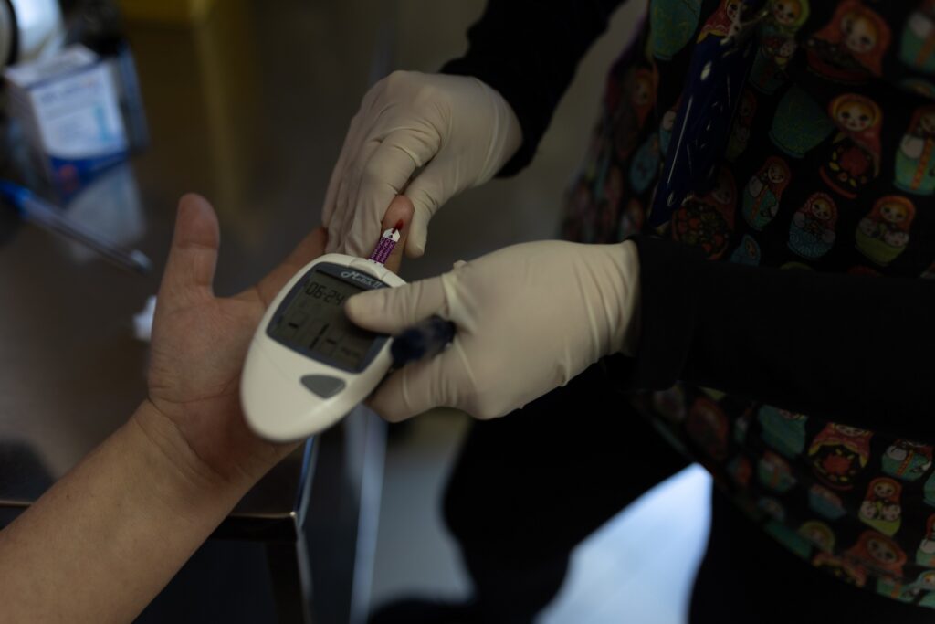 A imagem mostra um close-up de uma profissional de saúde usando um medidor de glicose para verificar os níveis de glicose no sangue de um paciente. A profissional de saúde está usando luvas e está cuidadosamente segurando o dedo do paciente para obter uma amostra de sangue. O medidor de glicose mostra uma leitura de 06:24. O fundo é desfocado, mas é possível ver que estão num ambiente clínico. A imagem transmite uma sensação de cuidado e atenção aos detalhes. A profissional de saúde parece estar focada em seu trabalho e comprometida em fornecer um serviço preciso e eficiente para o paciente. A imagem também destaca a importância de monitorar os níveis de glicose no sangue para pessoas com diabetes.