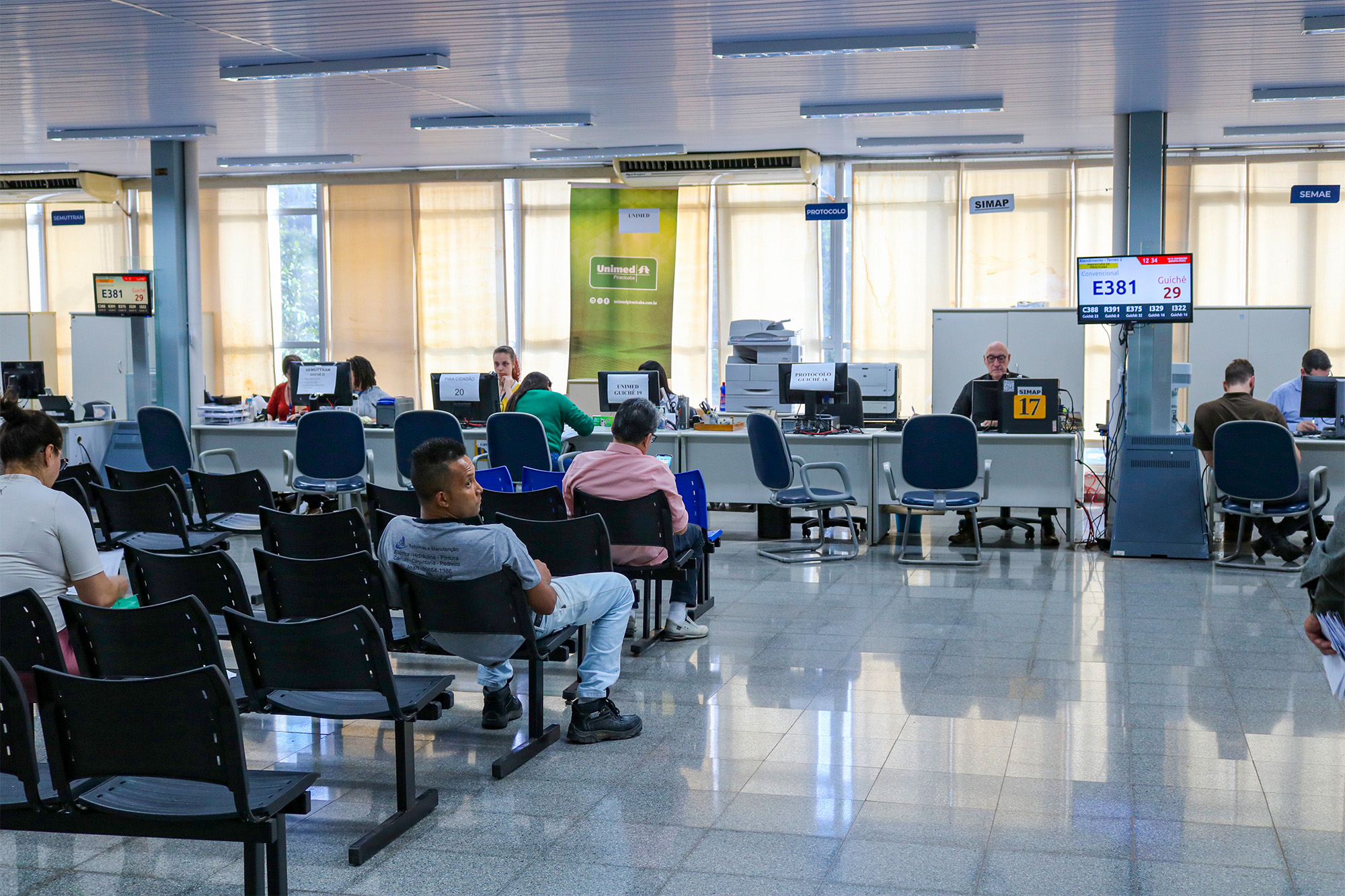 A imagem mostra uma sala de espera em um órgão público, possivelmente um posto de atendimento. Há várias filas de cadeiras pretas para os clientes esperarem. Atrás das cadeiras, há vários balcões com funcionários atendendo clientes em seus computadores. Há também displays eletrônicos exibindo números de guichê e senhas. As paredes são claras e as janelas são grandes e permitem a entrada de luz natural. A atmosfera é organizada e funcional. Há uma placa com o logotipo da Unimed visível.