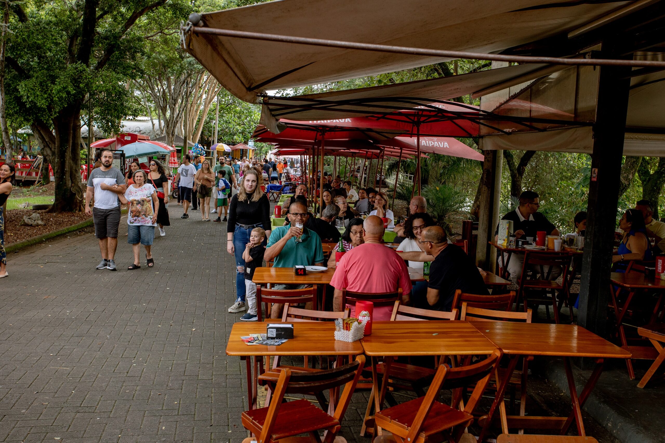 A imagem mostra uma cena movimentada em um restaurante ao ar livre, situado em uma área arborizada. Há diversas mesas de madeira com cadeiras de madeira, onde pessoas estão sentadas, conversando e comendo. A maioria está sob guarda-sóis vermelhos, com uma grande cobertura adicional protegendo uma área maior. O restaurante parece casual e aconchegante, com uma atmosfera familiar. As pessoas são de diferentes idades e parecem estar relaxadas e aproveitando o ambiente. Há pessoas caminhando ao fundo, sugerindo que o local é popular e parte de uma área mais ampla, talvez um parque ou praça. A iluminação é difusa, indicando provavelmente um dia nublado ou com sombra. O piso é de tijolos, típico de áreas externas pavimentadas. A imagem é rica em detalhes, mostrando a variedade de pessoas presentes, a disposição do mobiliário e o ambiente agradável do restaurante ao ar livre. A sensação geral é de um dia comum em um local popular e acolhedor.