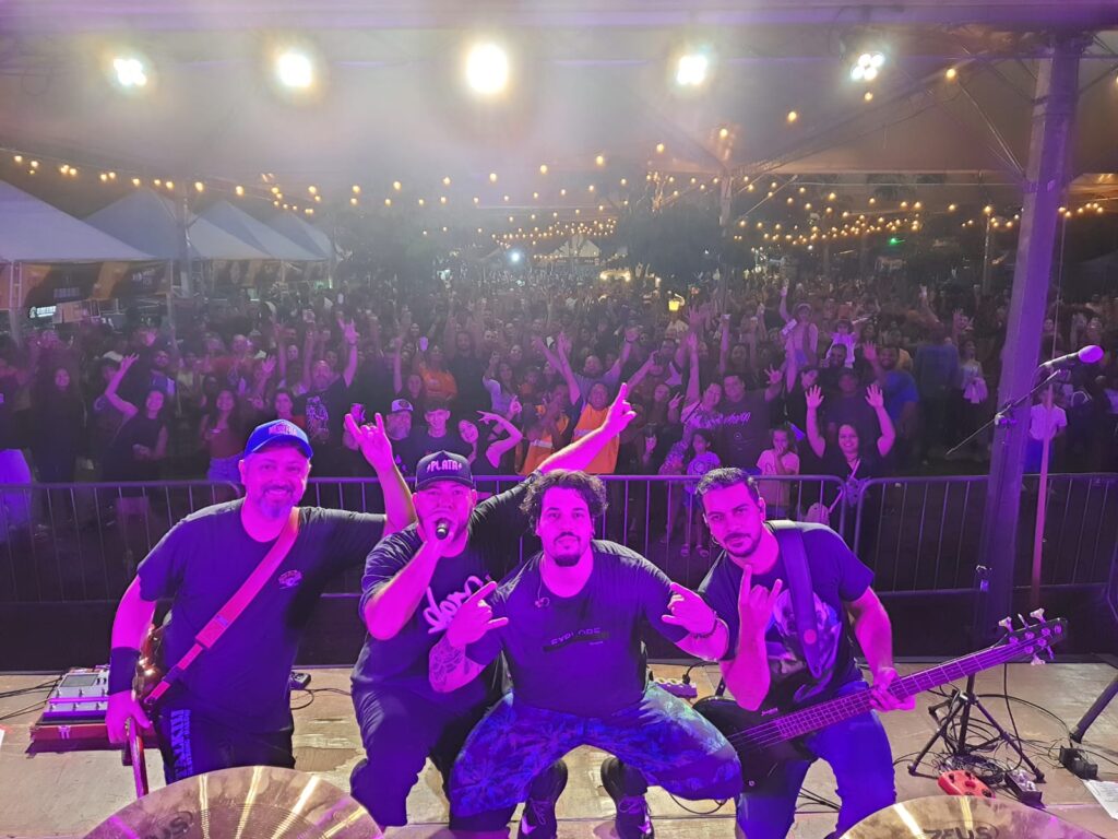 A imagem mostra uma banda de quatro membros se apresentando em um palco ao ar livre. Eles estão sorrindo para a câmera, e atrás deles há uma grande multidão de pessoas que estão animadas e com os braços levantados. A iluminação do palco é roxa e cria um ambiente vibrante. Os membros da banda estão usando roupas escuras e cada um parece ter seu próprio estilo único. A banda está posicionada de forma que o baixista e o vocalista central estão mais próximos da câmera. Há equipamentos de áudio visíveis no palco, como microfones, caixas de som e pratos de bateria. A multidão parece estar bastante próxima do palco, criando uma atmosfera íntima e energética. O local parece ser uma área aberta com tendas e iluminação externa. No geral, a imagem transmite a energia de um show ao vivo bem sucedido.
