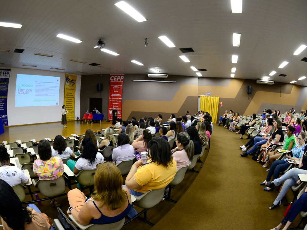 A imagem mostra um ambiente interno onde está ocorrendo uma apresentação ou palestra. Há um grande grupo de pessoas sentadas em fileiras, voltadas para a frente da sala, onde há uma tela exibindo algum texto. O público parece estar atento ao palestrante ou à apresentação. Na frente, há um pódio com microfones e banners de cada lado. A sala tem paredes amarelas e luzes montadas no teto.