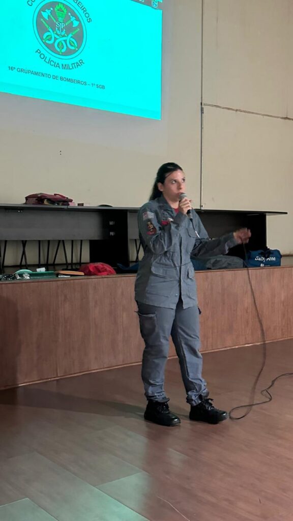 Karen Ribeiro Marques,cabo do Grupamento de Bombeiros de Piracicaba, com camisa e calça cinzas e botas pretas, fala ao microfone.