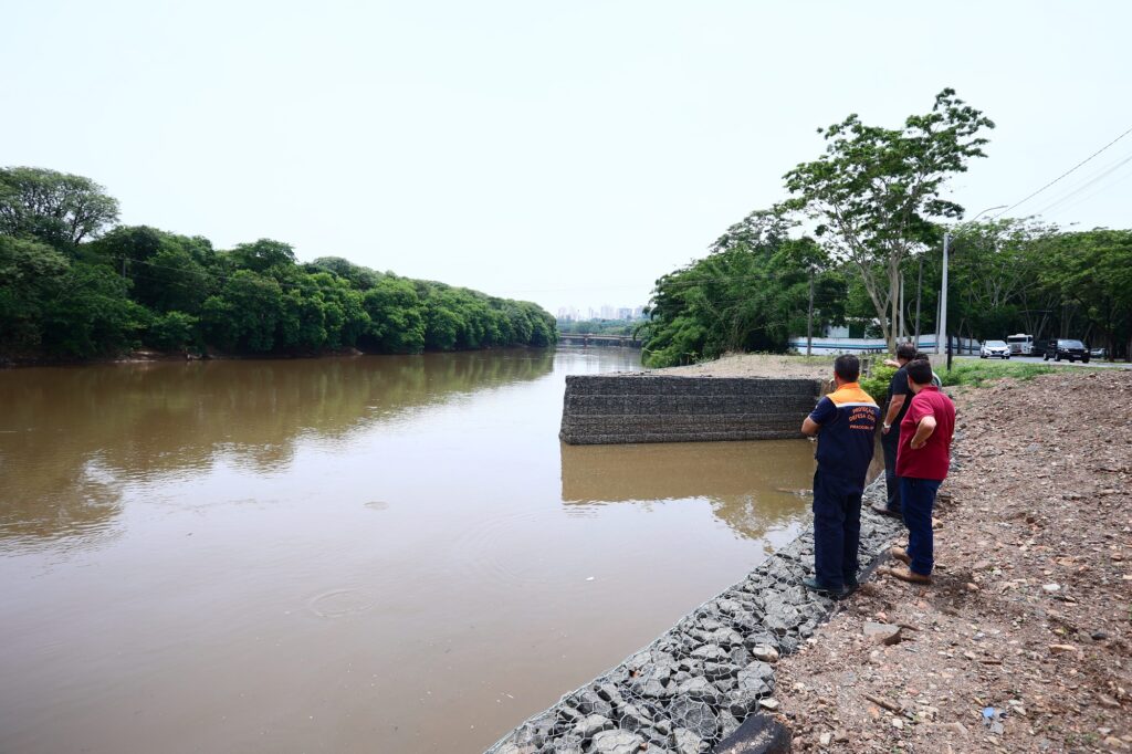 A imagem mostra um rio com água de cor marrom, margeado por árvores verdes e densas no lado esquerdo. À direita, há um talude rochoso com um muro de contenção feito de malha de arame e pedras. Três pessoas estão de pé no talude, olhando para o rio. Ao fundo, à direita, há uma estrada com vários veículos e mais árvores.