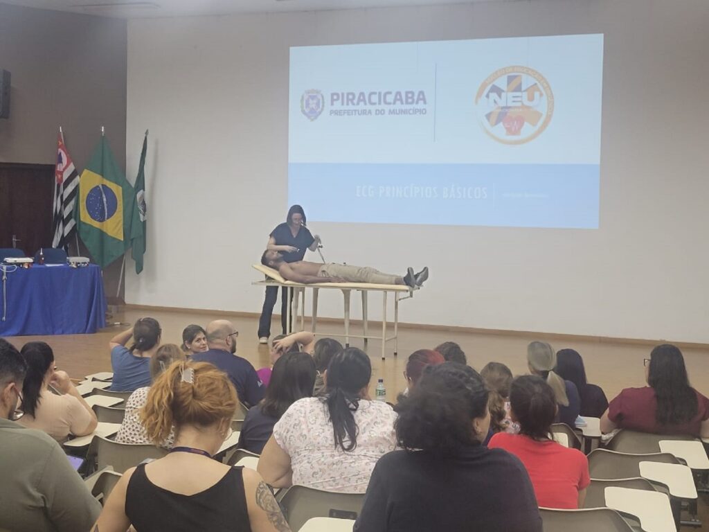 Mulher de roupa escura mexe em um paciente homem, para explicar os cuidados básicos, para platéia de enfermeiras.