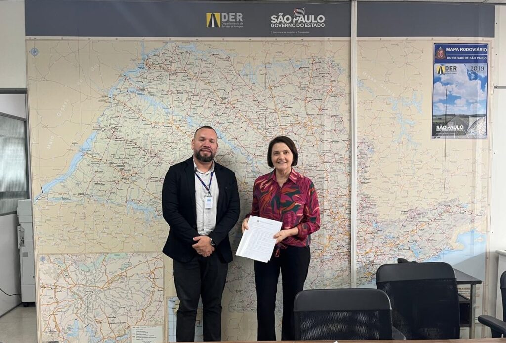 A imagem mostra duas pessoas em pé diante de um grande mapa do estado de São Paulo, Brasil. O mapa está rotulado com "DER Departamento de Estradas de Rodagem" e "São Paulo Governo do Estado". Uma das pessoas está segurando um documento. O mapa parece ser um mapa rodoviário detalhado de São Paulo, com várias rodovias e estradas marcadas. Há também um mapa menor no canto inferior esquerdo e um pôster do lado direito rotulado "Mapa Rodoviário do Estado de São Paulo 2019".
