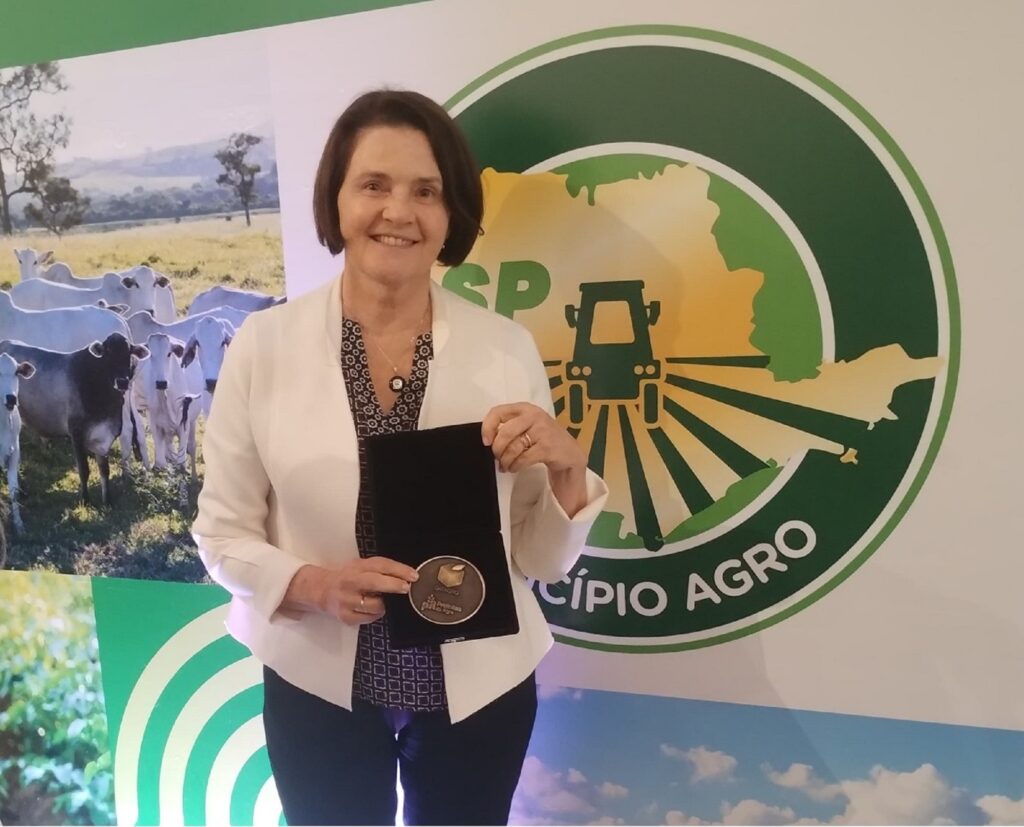 Secretaria municipal de Agricultura e Abastecimento (Sema), de blaiser claro e calça escura, segura medalha, tendo ao fundo o brasão de município agro