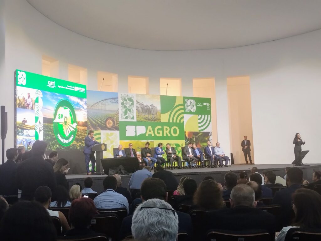 Evento do Programa Município Agro, do Governo do Estado de São Paulo. Ao microfone fala o secretário Estadual de Agricultura, tendo ao lado diversas autoridades sentadas e embaixo a platéia de pessoas que ouvem atentamente as explicações.