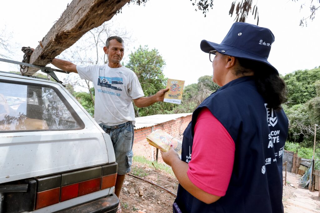 A imagem mostra um homem e uma mulher em uma interação ao ar livre. O homem, com idade aparente entre 40 e 50 anos, de pele bronzeada, usa uma camiseta branca com a inscrição "CADASTRE-SE" e shorts jeans. Ele está carregando uma grande viga de madeira em seu carro. O carro parece antigo e está estacionado ao lado de uma parede de tijolos. A mulher, que parece ser uma funcionária de saúde ou de serviço público, veste um boné azul-escuro, uma camiseta rosa e um colete azul-escuro com o logotipo de uma organização (provavelmente de saúde pública). Ela está entregando um folheto ao homem. O folheto contém ilustrações e o título "NÓS CONTRA O MOSQUITO". O fundo apresenta vegetação típica de clima tropical, com árvores e arbustos. O cenário parece ser uma área rural ou periférica. A cena captura um momento de interação entre uma funcionária de saúde pública e um morador, possivelmente durante uma campanha de conscientização sobre prevenção de doenças transmitidas por mosquitos, como a dengue ou Zika.
