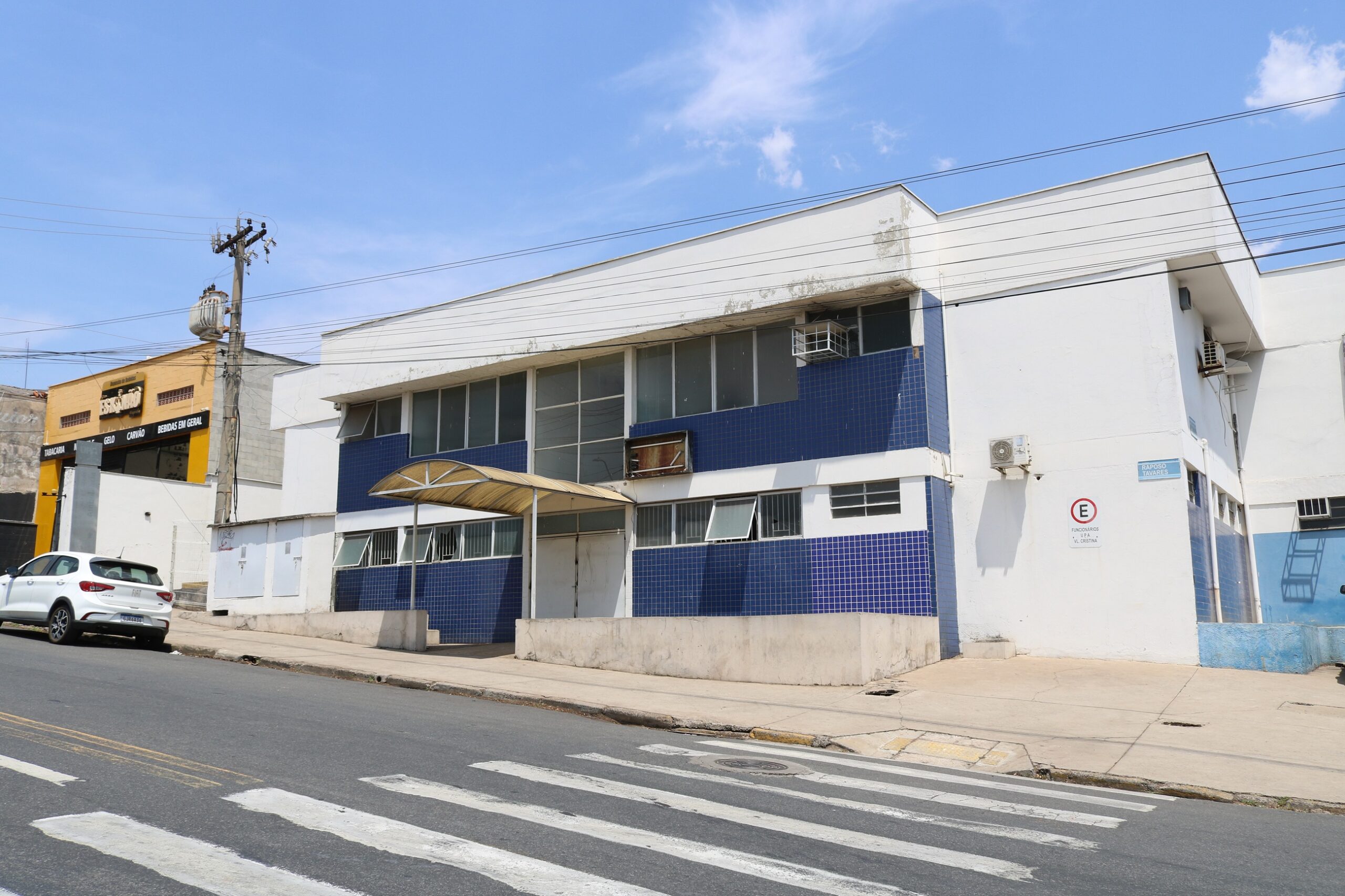 A imagem mostra um prédio de dois andares, de cor branca com detalhes em azul, localizado em uma rua. O prédio parece ser comercial ou institucional. À esquerda, há um prédio menor e amarelo, possivelmente um comércio. Na frente do prédio principal, há uma faixa de pedestre. O céu está azul e ensolarado. A imagem sugere uma área urbana, possivelmente em uma cidade brasileira, dado o estilo arquitetônico e a sinalização.