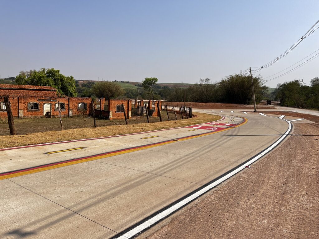 A imagem mostra uma estrada curva com marcações de faixa claras, incluindo linhas amarelas e brancas, e uma seção vermelha com símbolos brancos de bicicleta indicando uma ciclovia. No lado esquerdo da estrada, há uma estrutura de tijolos deteriorada com várias aberturas que parecem ser janelas e portas. A estrutura está cercada por uma cerca feita de postes de madeira e arame. Ao fundo, há árvores e uma paisagem montanhosa. O céu está claro e azul, sugerindo um dia ensolarado. Também há linhas de energia correndo paralelamente à estrada no lado direito.