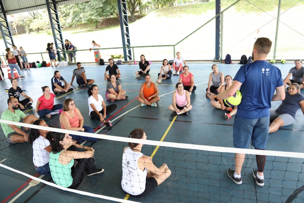 A imagem mostra um grupo de professores sentados no chão de uma quadra esportiva, participando de uma atividade. No centro, um treinador masculino está de pé, segurando uma bola e dando instruções aos participantes. O grupo é composto por adultos de diferentes idades e etnias, todos atentos ao que o treinador está dizendo. O ambiente é descontraído e inclusivo, refletindo um clima de amizade e colaboração. Ao fundo, é possível ver outras pessoas observando ou aguardando sua vez. A cena transmite uma energia positiva e ativa em torno do esporte adaptado.