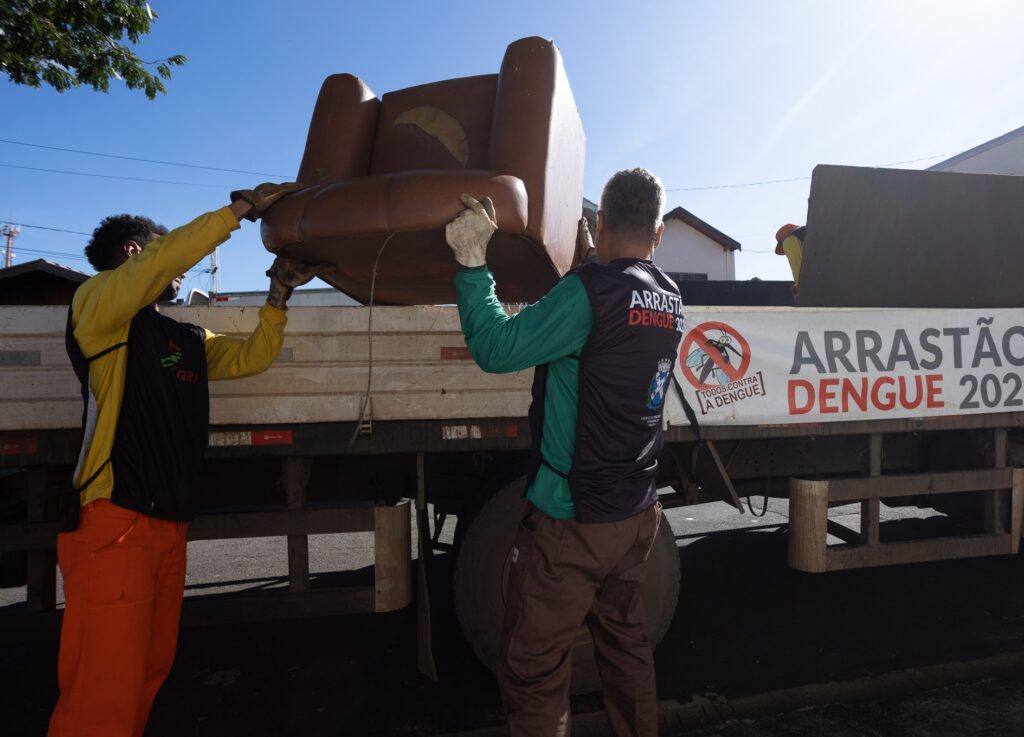A foto mostra dois homens carregando um sofá velho para a carroceria de um caminhão. O caminhão tem um letreiro que diz "Arrastão Dengue 202[ano ilegível]", indicando que é parte de uma campanha de saúde pública para combater a dengue. Um dos homens veste uma camisa amarela e calças laranja, enquanto o outro veste uma camisa verde e calças marrom. Ambos usam luvas. Ao fundo, há casas e árvores, e o céu está claro e azul. A cena sugere um esforço comunitário para remover objetos que podem acumular água e servir como criadouros para mosquitos transmissores da dengue.