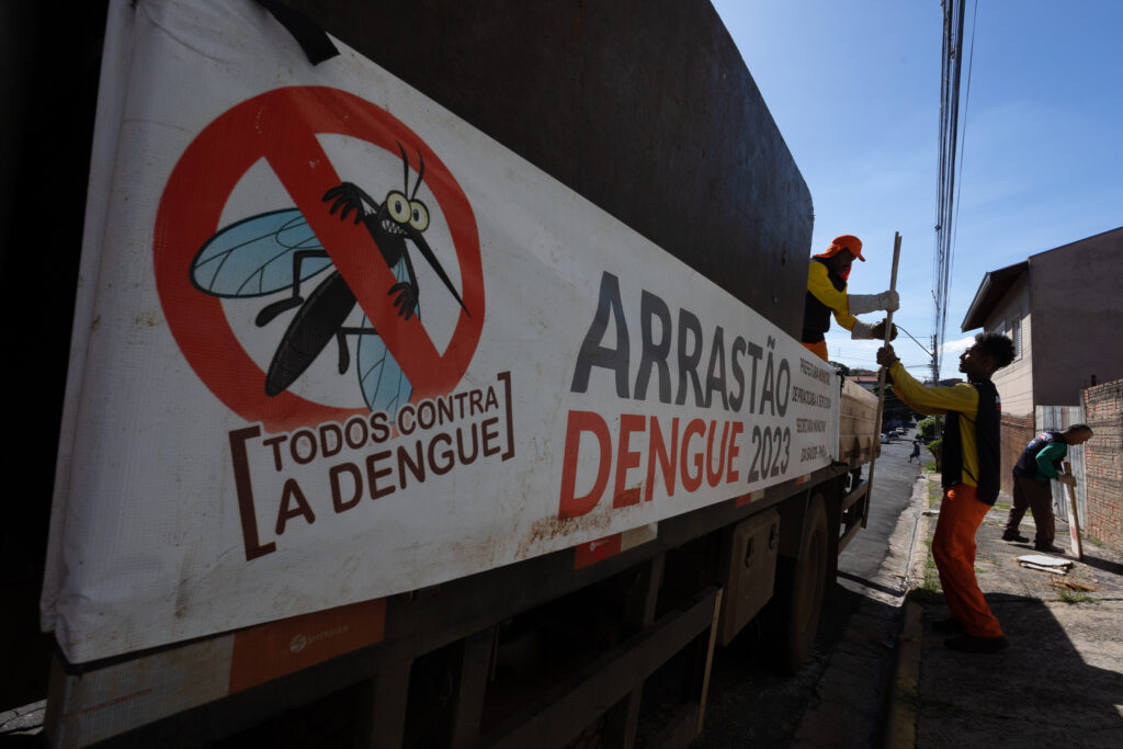 A imagem mostra um caminhão com uma faixa que diz "Arrastão Dengue 2023" e "[Todos contra a dengue]". Há um desenho de um mosquito com um círculo vermelho de proibição sobre ele. Três pessoas estão carregando objetos no caminhão. A cena parece ser uma ação de limpeza pública para combater a dengue.