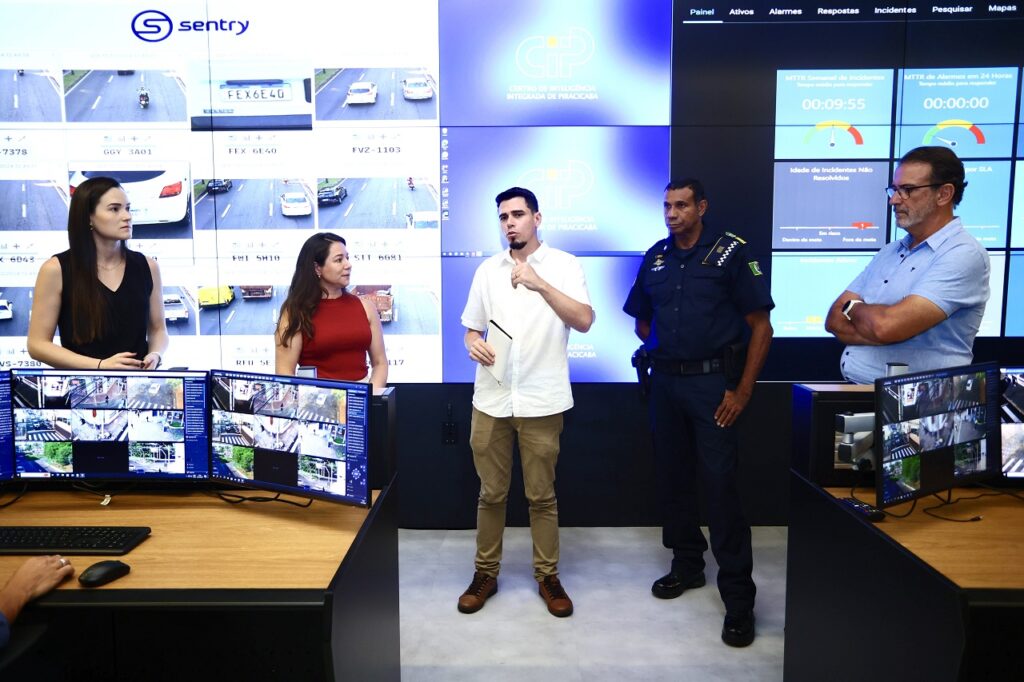A imagem mostra uma sala de controle de monitoramento de segurança, com várias telas exibindo imagens de câmeras de segurança espalhadas por uma cidade. Há vários funcionários em frente às telas, possivelmente monitorando a atividade e respondendo a incidentes. A sala parece moderna e tecnológica, sugerindo uma operação de segurança sofisticada e eficiente. A imagem também mostra dados estatísticos de incidentes, incluindo o tempo médio para resolução.