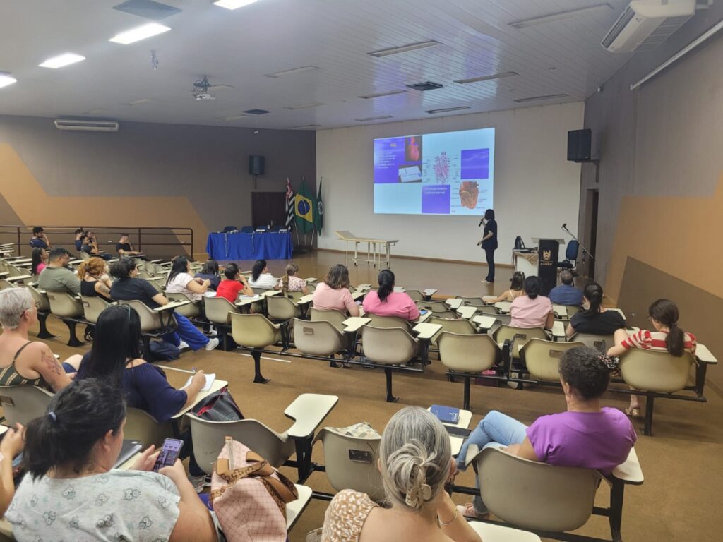 Mulher com roupa escura e com microfone na mão, olha para um telão com diversas imagens, para uma platéia formada por enfermeiras, que estão devidamente sentadas.