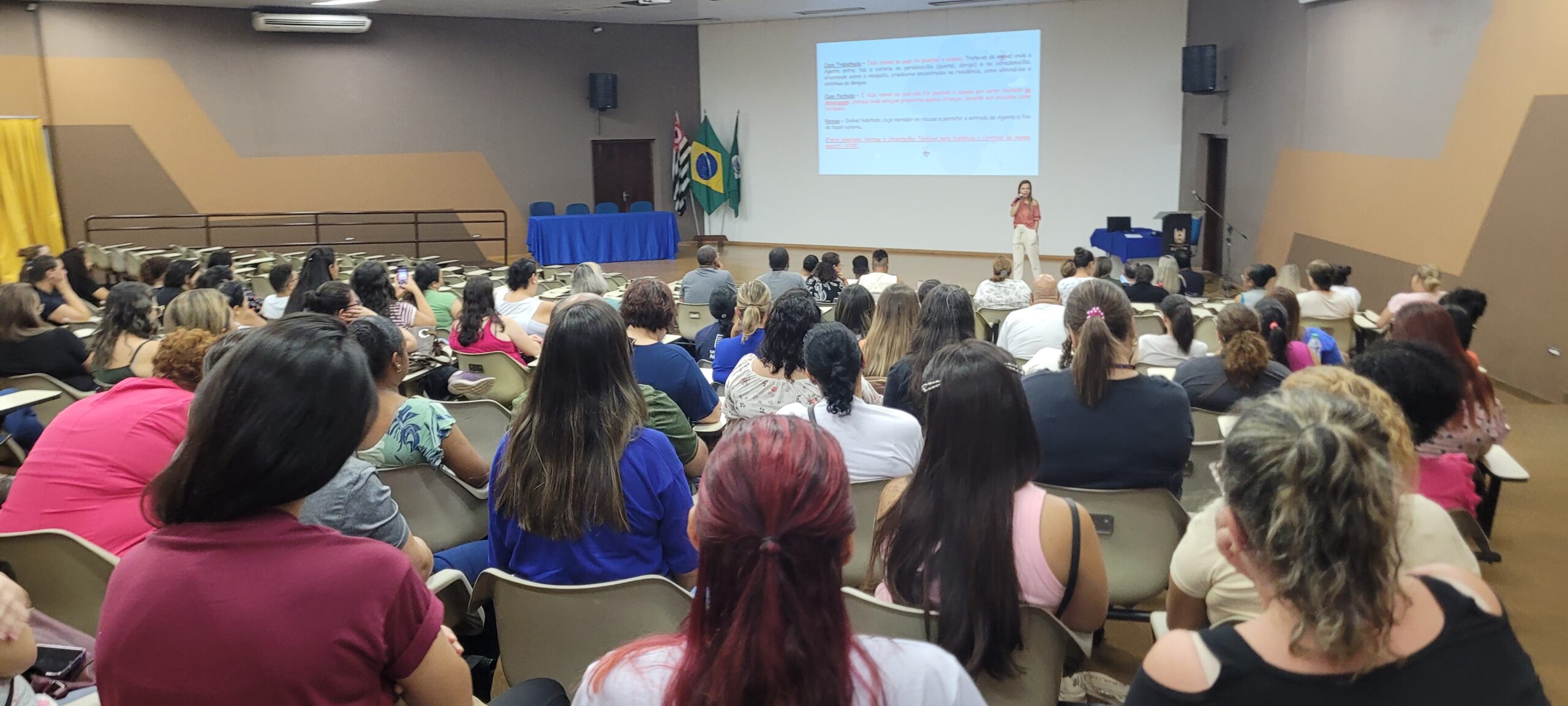 A imagem mostra uma sala de aula lotada de mulheres sentadas em fileiras de cadeiras. Uma palestrante está em pé no palco, dando uma apresentação. No palco, há um projetor mostrando uma apresentação de slides. A sala de aula é bem iluminada e há bandeiras brasileiras penduradas na parede. A maioria das mulheres está vestida com roupas casuais. A atmosfera é calma e atenciosa. Parece ser uma reunião ou uma palestra.