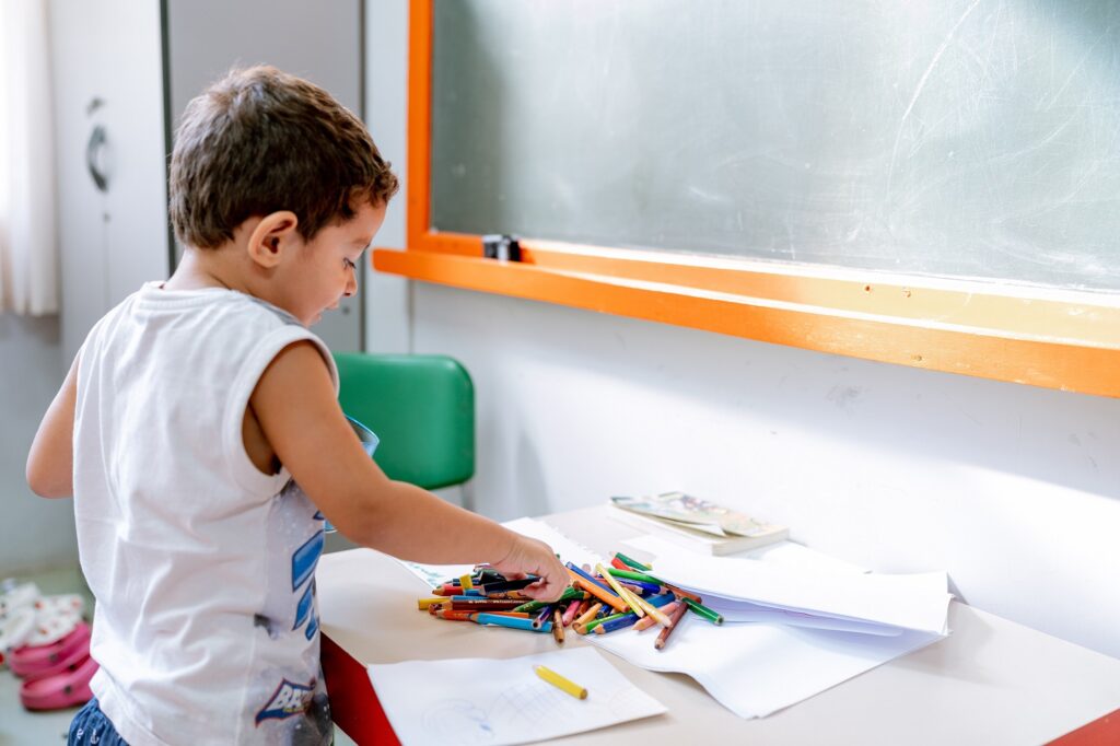 Criança com camiseta branca brinca com lápis coloridos espalhados em uma mesinha branca. Ele está sorrindo e olhando atentamente para os objetos.