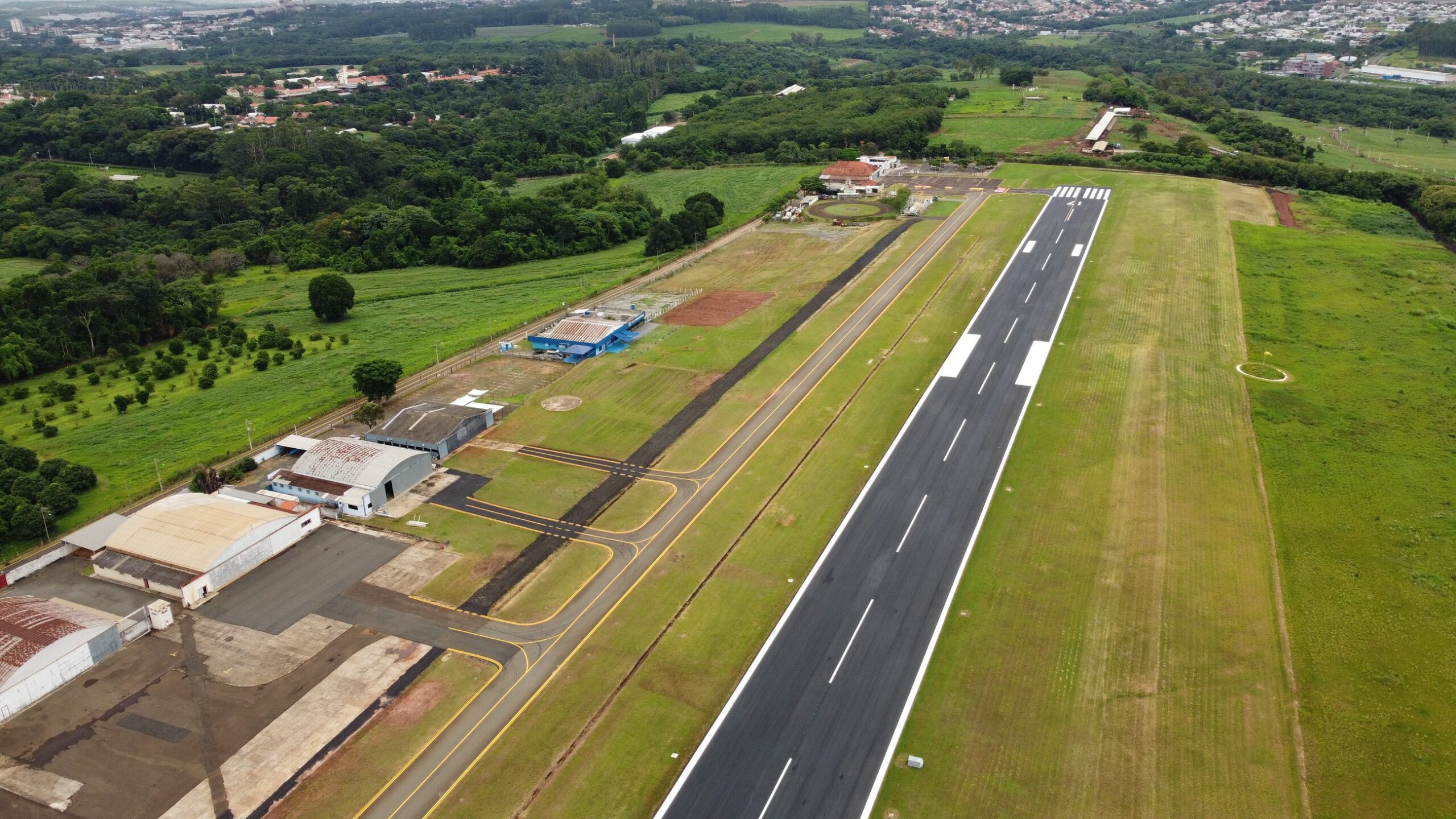 A imagem mostra uma vista aérea de um pequeno aeroporto. A pista de pouso e decolagem é o elemento central, com marcações claras e linhas de demarcação. Ao redor da pista, há áreas gramadas, hangares de diferentes tamanhos e outras estruturas associadas a um aeroporto. Há também um campo aberto, possivelmente uma área de estacionamento ou manutenção. A vegetação circundante é predominantemente verde, sugerindo uma área rural ou suburbana. A imagem oferece uma perspectiva geral da infraestrutura e do layout do aeroporto, mostrando sua dimensão e as instalações existentes.