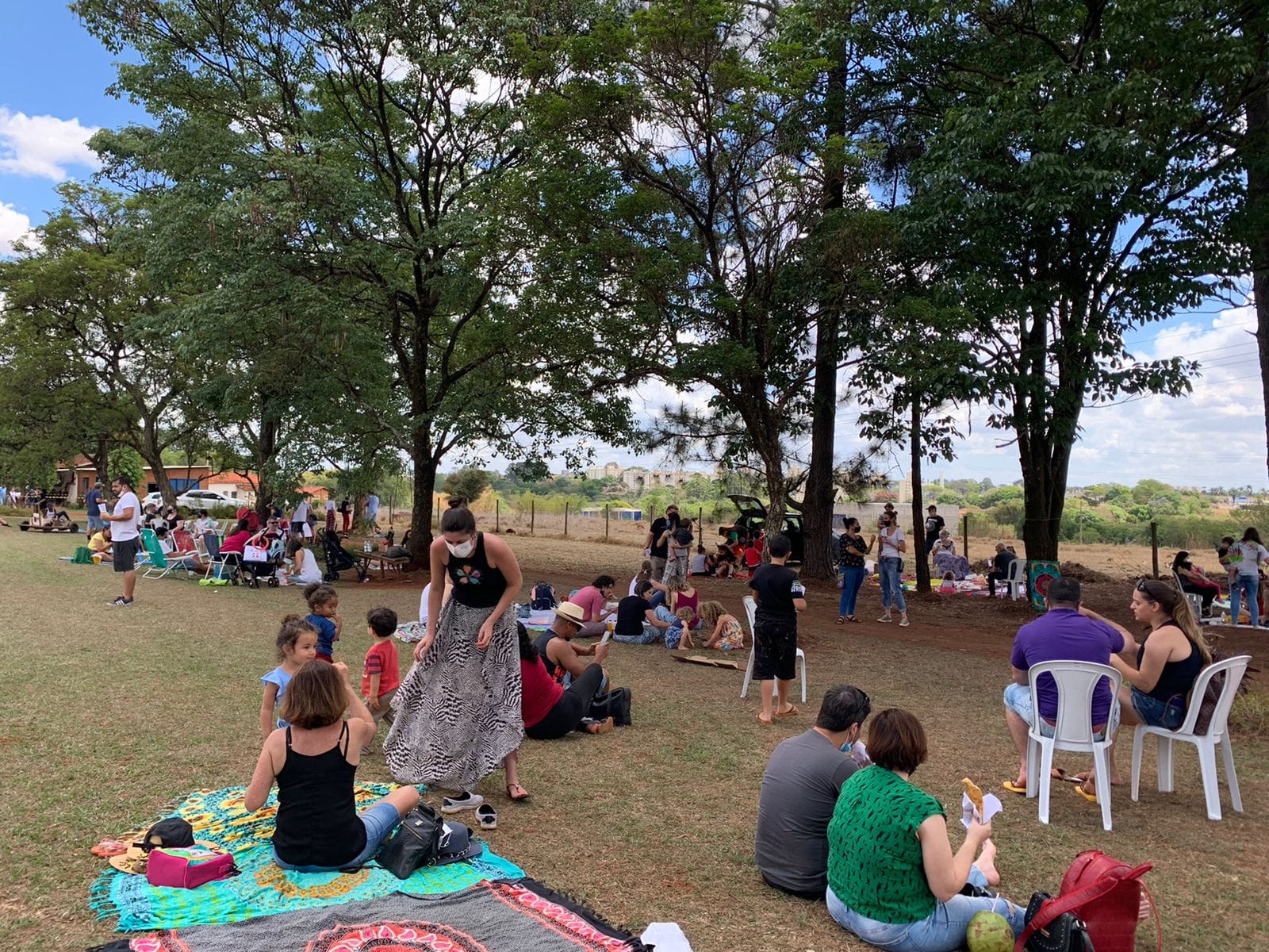 A imagem mostra um grande grupo de pessoas reunidas em um piquenique em um gramado gramado sob a sombra de árvores altas. Muitas famílias estão presentes, com crianças brincando e adultos sentados em mantas de piquenique. Há cadeiras de plástico espalhadas pela área, e algumas pessoas usam máscaras faciais. No fundo, há uma linha de árvores e alguns prédios visíveis à distância. O ambiente parece ser um parque ou área de lazer ao ar livre. A atmosfera geral é de um evento comunitário ou encontro familiar casual. A imagem está bem iluminada, parecendo ter sido tirada em um dia ensolarado.