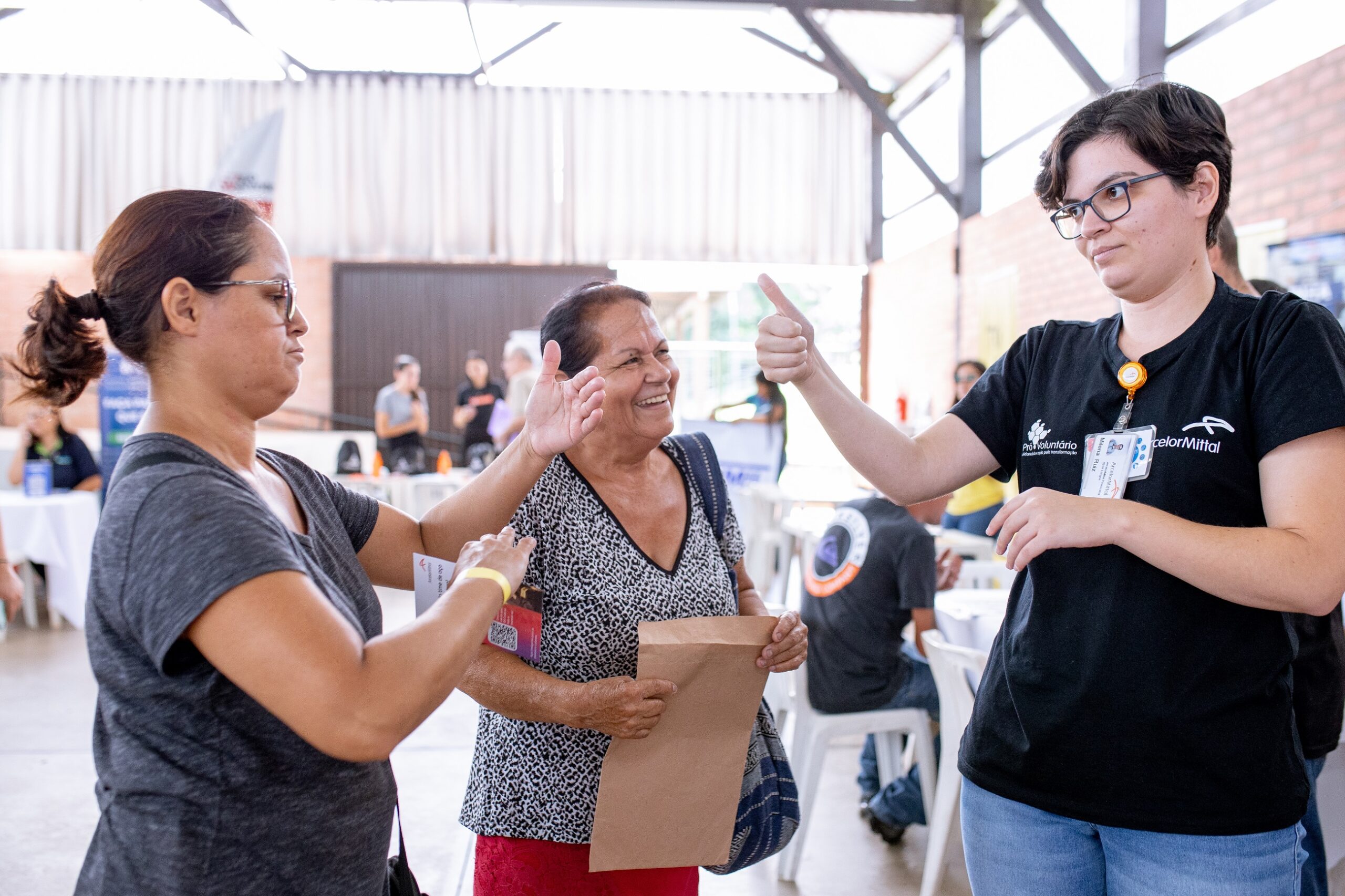 A imagem mostra três mulheres em uma sala grande e bem iluminada, possivelmente um ginásio ou centro comunitário. Uma mulher mais velha, de pele morena e cabelos curtos escuros, está no centro, sorrindo. Ela está vestida com uma blusa estampada em preto e branco e uma saia vermelha. Ela segura um envelope pardo. À esquerda dela, uma mulher mais jovem, também morena, com cabelos castanhos presos em um rabo de cavalo, está com o braço ao redor da mulher mais velha, a abraçando. Ela parece estar ajudando ou interagindo com a mulher mais velha. Está vestindo uma camiseta cinza escura. À direita, uma mulher mais jovem com óculos e cabelo curto escuro está dando um "joinha" para a mulher mais velha, e a outra mulher parece estar lhe entregando algo. Esta mulher está usando uma camisa preta com o logotipo de uma organização e um crachá de identificação visível. Ela parece ser uma voluntária ou funcionária do local. Ao fundo, há outras pessoas, mais desfocadas, sugerindo um evento ou reunião comunitária. Há mesas e cadeiras espalhadas pela sala. A atmosfera geral é amigável e acolhedora.