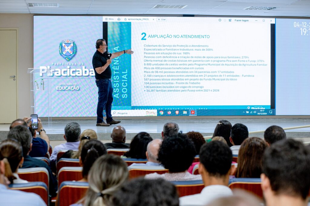 A imagem mostra o prefeito Luciano Almeida fazendo a apresentação em frente a uma audiência. A apresentação parece estar relacionada à assistência social e à educação, como indicado pelo texto na tela e no fundo. O fundo tem o logotipo e o nome "Prefeitura de Piracicaba" e "Secretaria de Educação". A tela exibe um slide intitulado "AMPLIAÇÃO NO ATENDIMENTO", que enumera várias estatísticas e realizações relacionadas aos serviços de assistência social.
