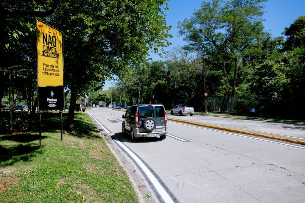 A imagem mostra uma cena de rua pavimentada com concreto com uma placa de trânsito proeminente no lado esquerdo. A placa diz: "NÃO COMETA IMPRUDÊNCIAS Preze pela vida. A paz no trânsito começa por você". A placa é um aviso para motoristas evitarem comportamentos imprudentes e enfatiza a importância de valorizar a vida e promover a paz no trânsito. A rua está cercada por árvores e tem alguns veículos, incluindo um SUV cinza e uma caminhonete branca.