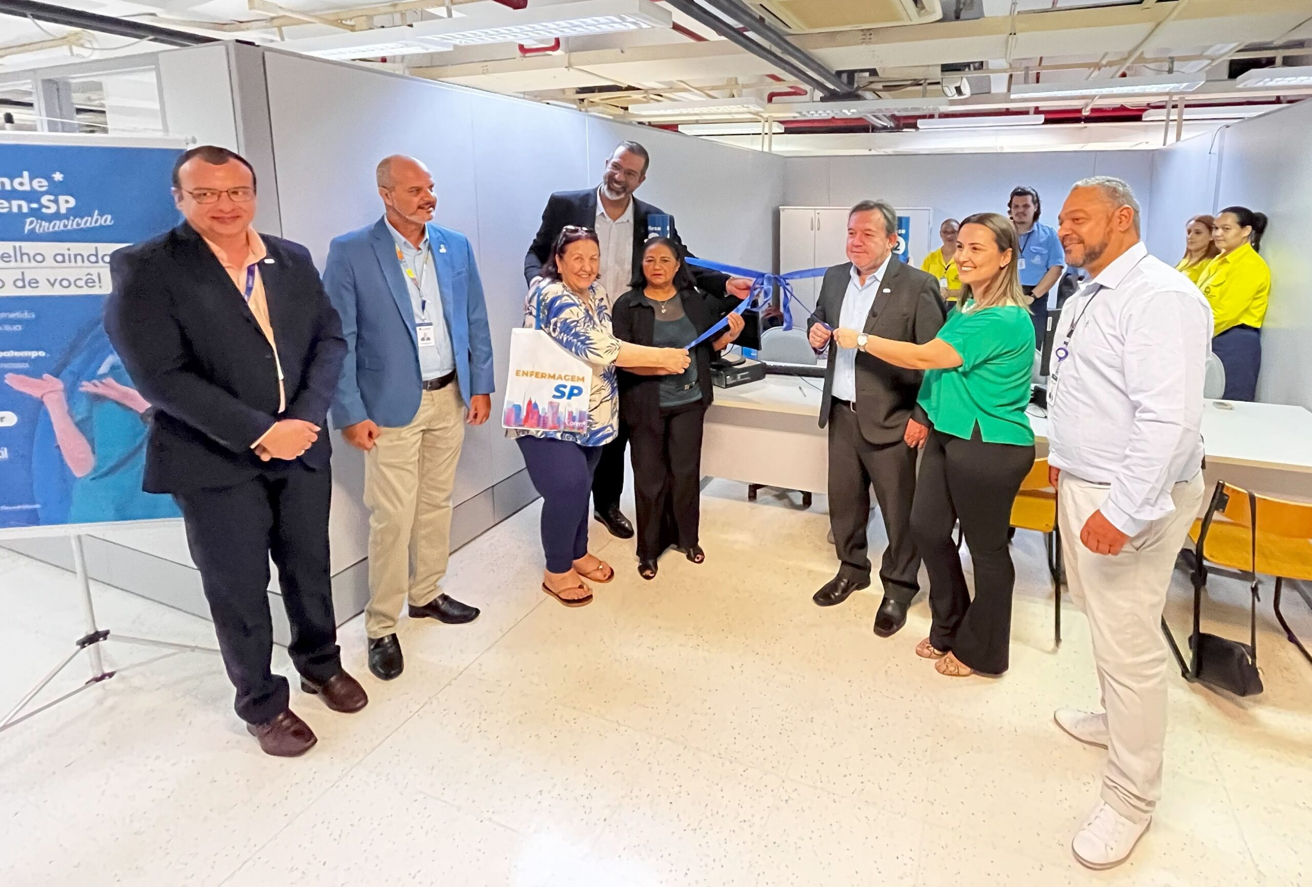 A imagem mostra um grupo de pessoas em um evento de inauguração. Há um laço azul sendo cortado por duas mulheres. Há um banner na parte esquerda da imagem com o logotipo do evento. As pessoas na imagem estão vestindo roupas formais. O ambiente parece ser um escritório ou uma sala de conferências.