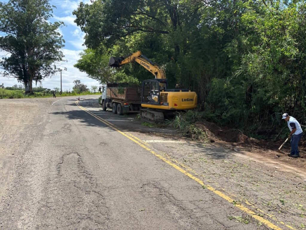 Caminhão com elevatória na cor amarela, no acostamento, fazendo a limpeza.Ao lado apareceu um homem de camiseta branca e calça escura, com um aenxada puxando a terra do local.