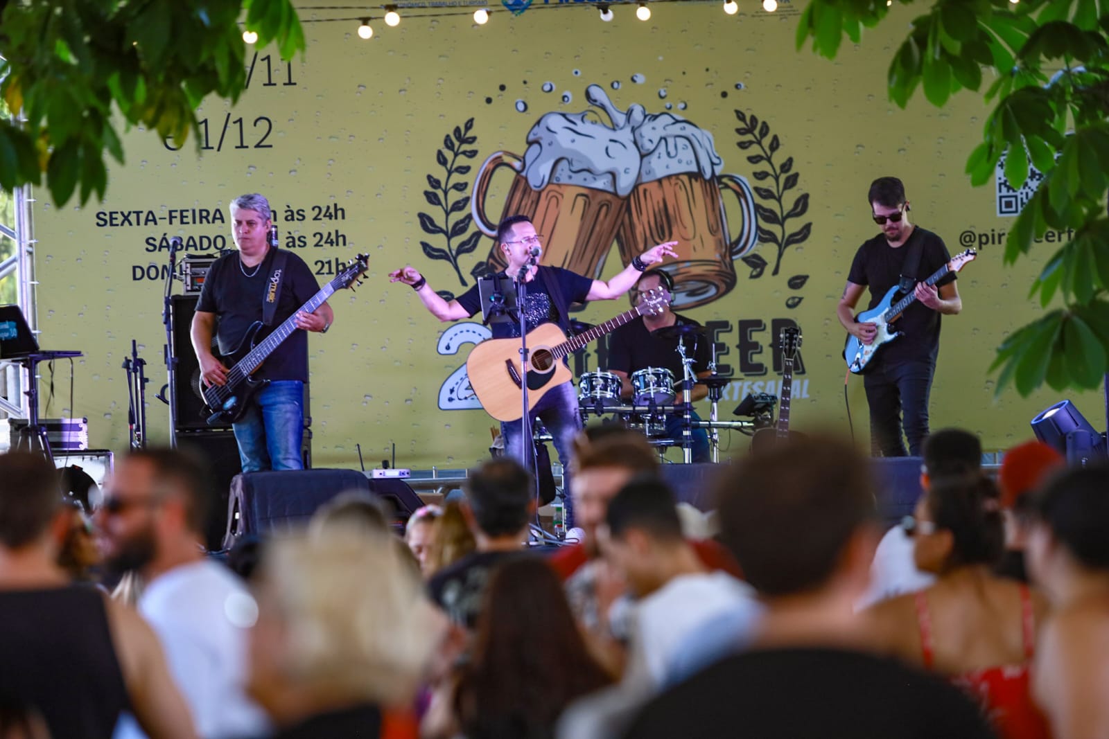 A imagem mostra uma banda tocando em um palco ao ar livre em frente a uma multidão. O palco tem um banner atrás dele com o título "2ª Beer Festival" e informações sobre os horários de funcionamento (sexta e sábado às 24h, domingo às 21h). A banda é composta por três músicos: um vocalista com guitarra acústica, um baixista e um guitarrista. O vocalista está no centro, com uma expressão animada enquanto canta e toca sua guitarra. O baixista está à sua esquerda, tocando um baixo preto. O guitarrista está à direita, tocando uma guitarra elétrica azul. A multidão na frente do palco é grande e composta por muitas pessoas, principalmente jovens adultos. As pessoas estão em pé e sentadas, algumas com bebidas na mão, outras apenas observando a banda. A atmosfera parece festiva e animada. Há uma iluminação suave no palco e no público, criando um ambiente aconchegante. O ambiente ao redor do palco sugere um espaço aberto, talvez um jardim ou praça, com árvores e luzes decorativas penduradas acima do palco. O foco principal da imagem é a banda e a energia da apresentação, mas a multidão animada contribui para a atmosfera geral.