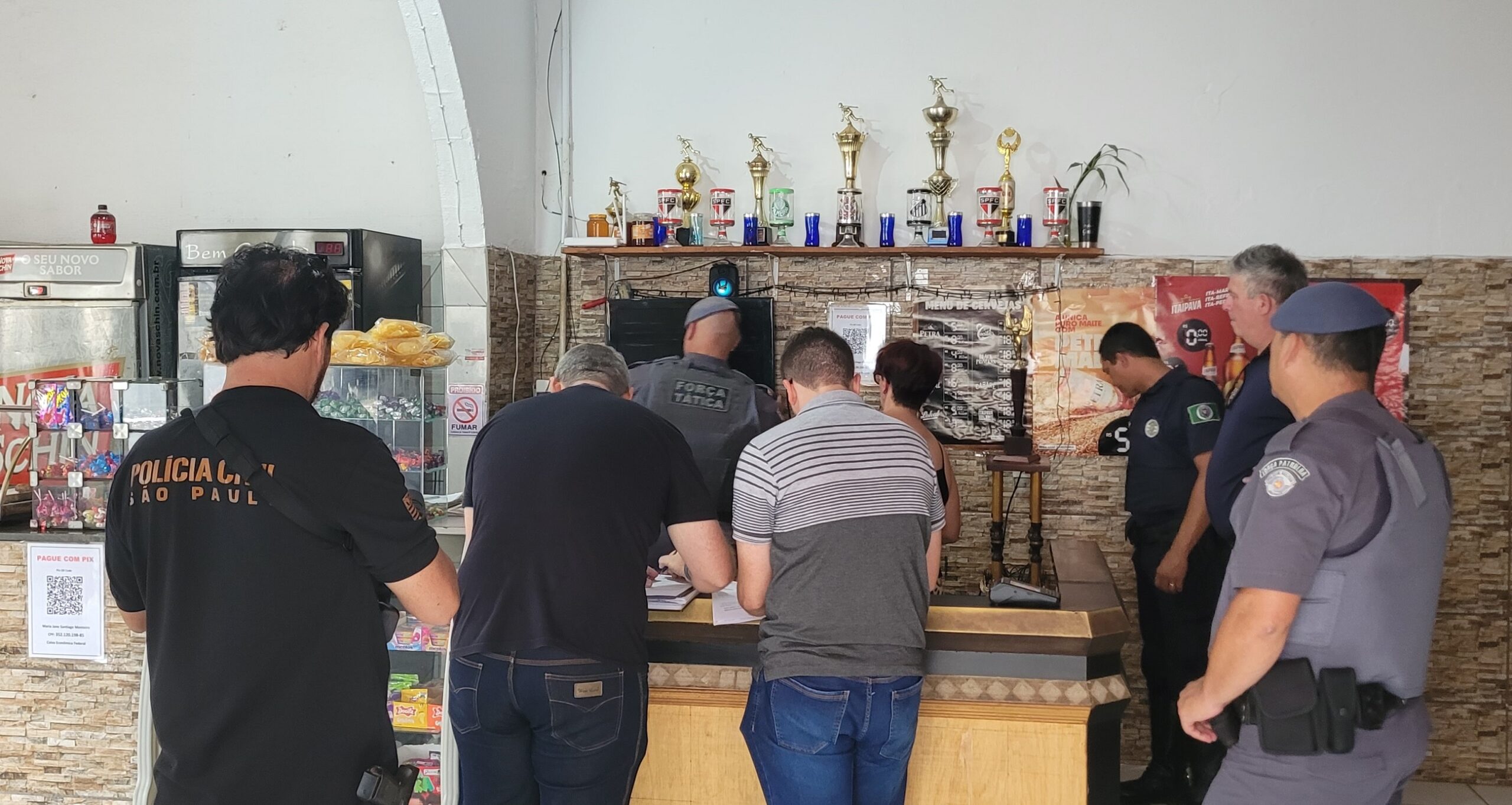 A imagem mostra vários policiais militares em uma ação policial em um estabelecimento comercial. Os policiais estão interagindo com os funcionários ou clientes do local, possivelmente realizando uma averiguação ou busca. Há uma grande quantidade de troféus e prêmios esportivos atrás do balcão, sugerindo que o lugar pode ser um bar ou restaurante que apoia times ou esportes locais. A ação parece ocorrer durante o dia. Há um cartaz com a logo da Polícia Militar do Estado de São Paulo em uma das camisetas dos policiais.