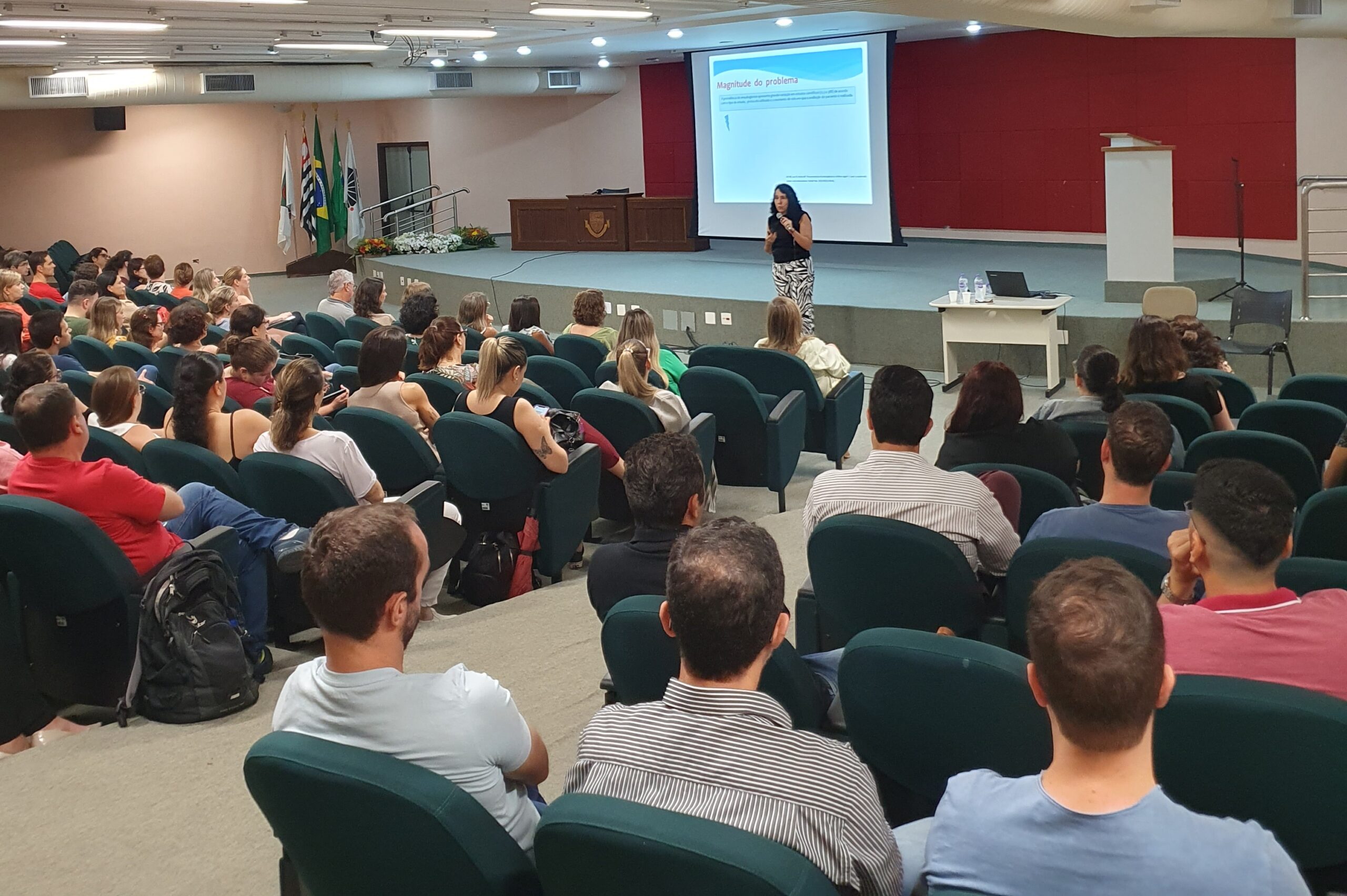 A imagem mostra uma palestra ou apresentação em um auditório. Uma mulher está no palco, de frente para a plateia, apresentando algo em um projetor. A tela do projetor mostra o título "Magnitude do problema". A plateia é composta por muitas pessoas sentadas em cadeiras de auditório verdes. As pessoas estão sentadas em fileiras, voltadas para o palco. A sala é bem iluminada e as pessoas parecem estar prestando atenção à apresentação. Há uma mesa com uma garrafa de água e um laptop no palco, ao lado da palestrante. No fundo, há bandeiras e um ambiente que parece ser uma universidade ou instituição de ensino. A imagem mostra uma grande quantidade de pessoas, e o foco é na palestrante e na plateia. As pessoas estão vestidas com roupas casuais, e a atmosfera parece formal, mas também acolhedora e atenciosa.