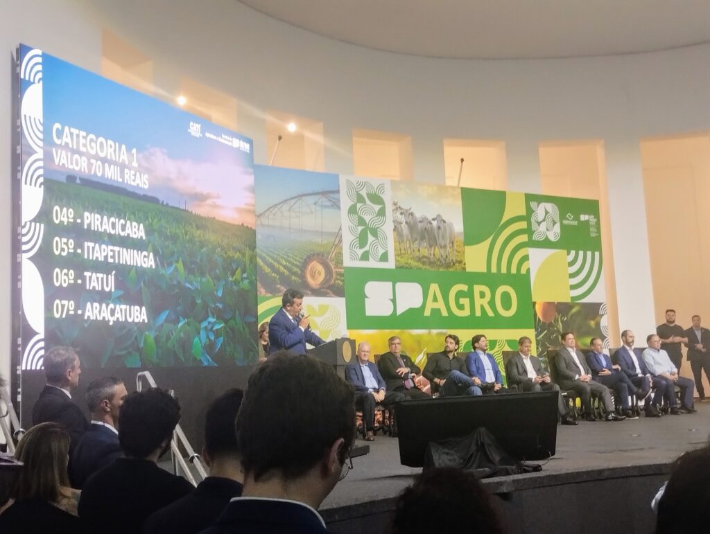 No telão do evento Município Agro em São Paulo, aparece o nome de Piracicaba em quarto lugar na premiação. O secretário estadual de Agricultura fala ao microfone, tendo ao lado nove autoridades sentadas.