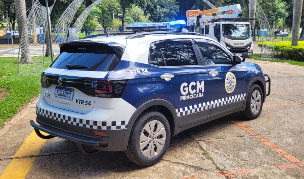 Esta é uma foto de uma viatura da Guarda Civil Municipal (GCM) de Piracicaba, São Paulo, Brasil. O veículo é uma Volkswagen T-Cross, de cor azul e branca, com a identificação da GCM e o número da viatura (VTR 54) visíveis. A placa do veículo também está visível. A foto foi tirada em uma área externa, possivelmente em um estacionamento ou pátio. Ao fundo, é possível ver um caminhão e uma cerca de arame farpado.