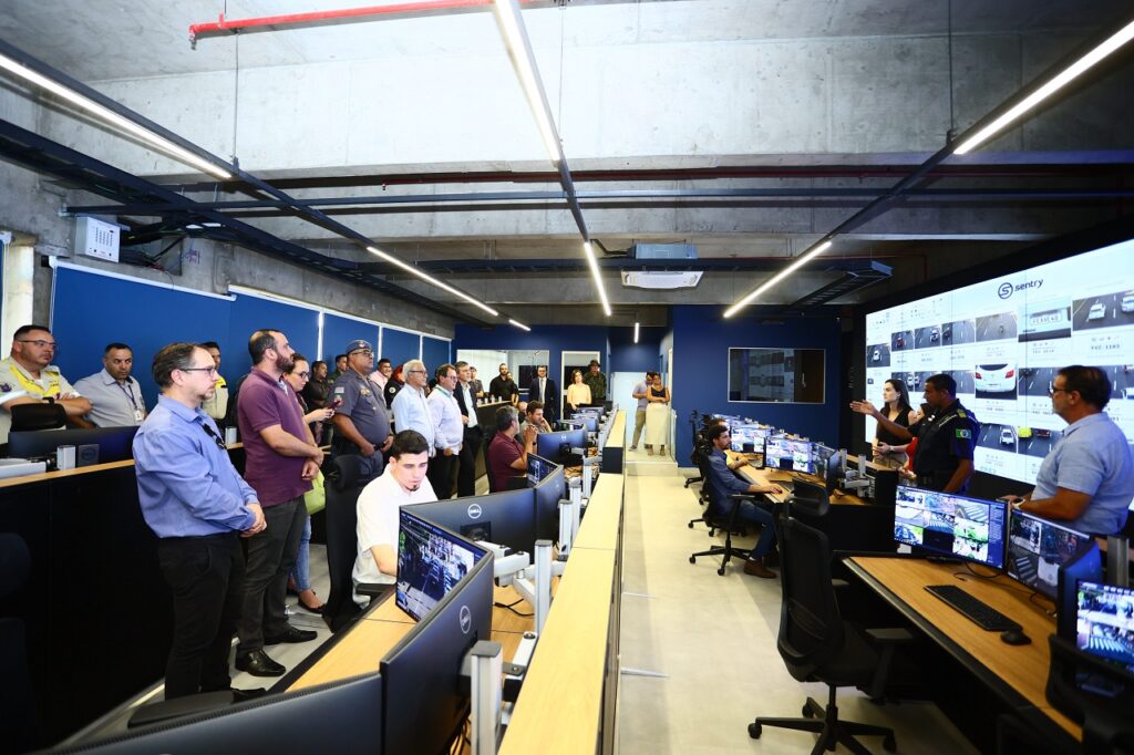 A imagem mostra uma sala de controle moderna e espaçosa, provavelmente usada para monitoramento de segurança ou tráfego. Há vários monitores de computador mostrando múltiplos feeds de vídeo, sugerindo um sistema de vigilância de circuito fechado (CCTV) extensivo.  A sala está cheia de pessoas, uma mistura de funcionários e possivelmente visitantes ou autoridades, observando os monitores ou participando de uma apresentação.  O design da sala é clean e funcional, com iluminação de teto moderna e um esquema de cores sóbrio. A atmosfera sugere uma atmosfera profissional e de alta tecnologia. Os funcionários presentes parecem estar de uniforme ou em trajes formais, indicando um ambiente corporativo ou governamental. Há um grande monitor na parede que mostra várias imagens de câmeras diferentes, dando uma visão geral do sistema de monitoramento.