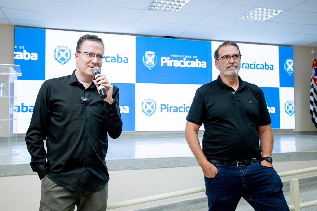 A imagem mostra Sérgio Pacheco e Luciano Almeida de pé em frente a um fundo que exibe o logotipo e o nome da "Prefeitura de Piracicaba". Sérgio está segurando um microfone e parece estar falando, enquanto Luciano está com uma das mãos no bolso. O fundo inclui o brasão de armas e o texto "Prefeitura de Piracicaba" repetido várias vezes. Há também uma bandeira visível no lado direito da imagem.