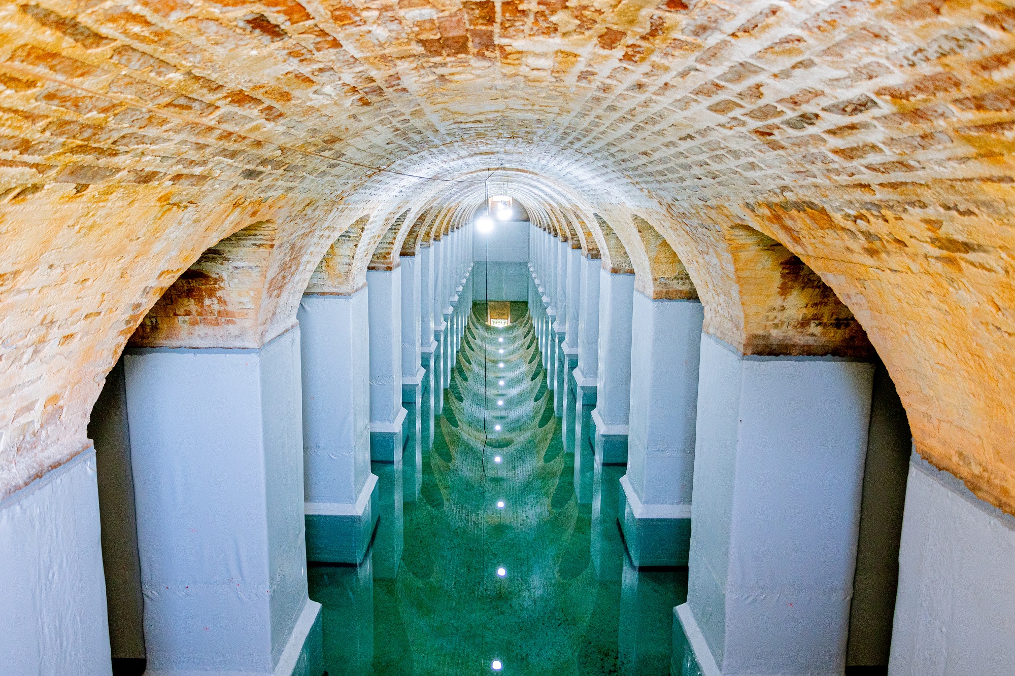 A imagem mostra um longo corredor estreito com uma série de arcos e colunas. O teto é de tijolo e tem uma série de luzes ao longo de sua extensão. As paredes e colunas são pintadas de branco e há uma superfície reflexiva, possivelmente água, no chão, que cria uma imagem espelhada dos arcos e luzes. No final do corredor, há uma pequena janela com uma grade decorativa, permitindo que alguma luz natural entre.