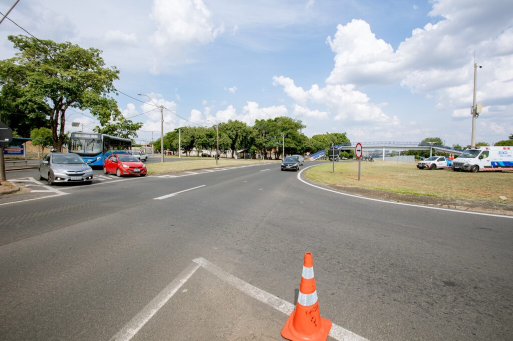 A imagem mostra um cruzamento de estradas com vários veículos, incluindo um ônibus, carros e vans. Há um cone de trânsito em primeiro plano e uma placa de "proibido entrar" no lado direito da estrada. O cruzamento é rodeado por árvores e áreas gramadas, com uma passarela de pedestres visível ao fundo. O céu está parcialmente nublado.