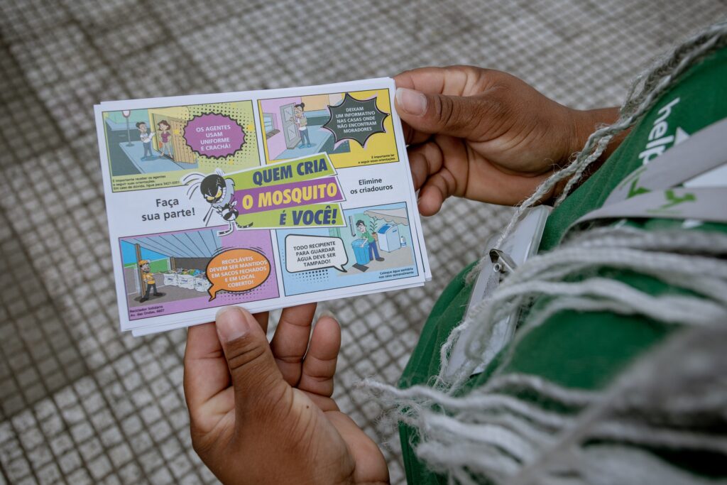 A imagem mostra uma pessoa segurando um folheto informativo sobre a prevenção da dengue. O folheto é um gibi que explica quem cria o mosquito e como evitar a proliferação dele. As instruções incluem usar uniforme e crachá, eliminar criadouros, manter recipientes tampados e descartar recicláveis em sacos fechados e em local coberto. O folheto também fornece um número de telefone para mais informações.