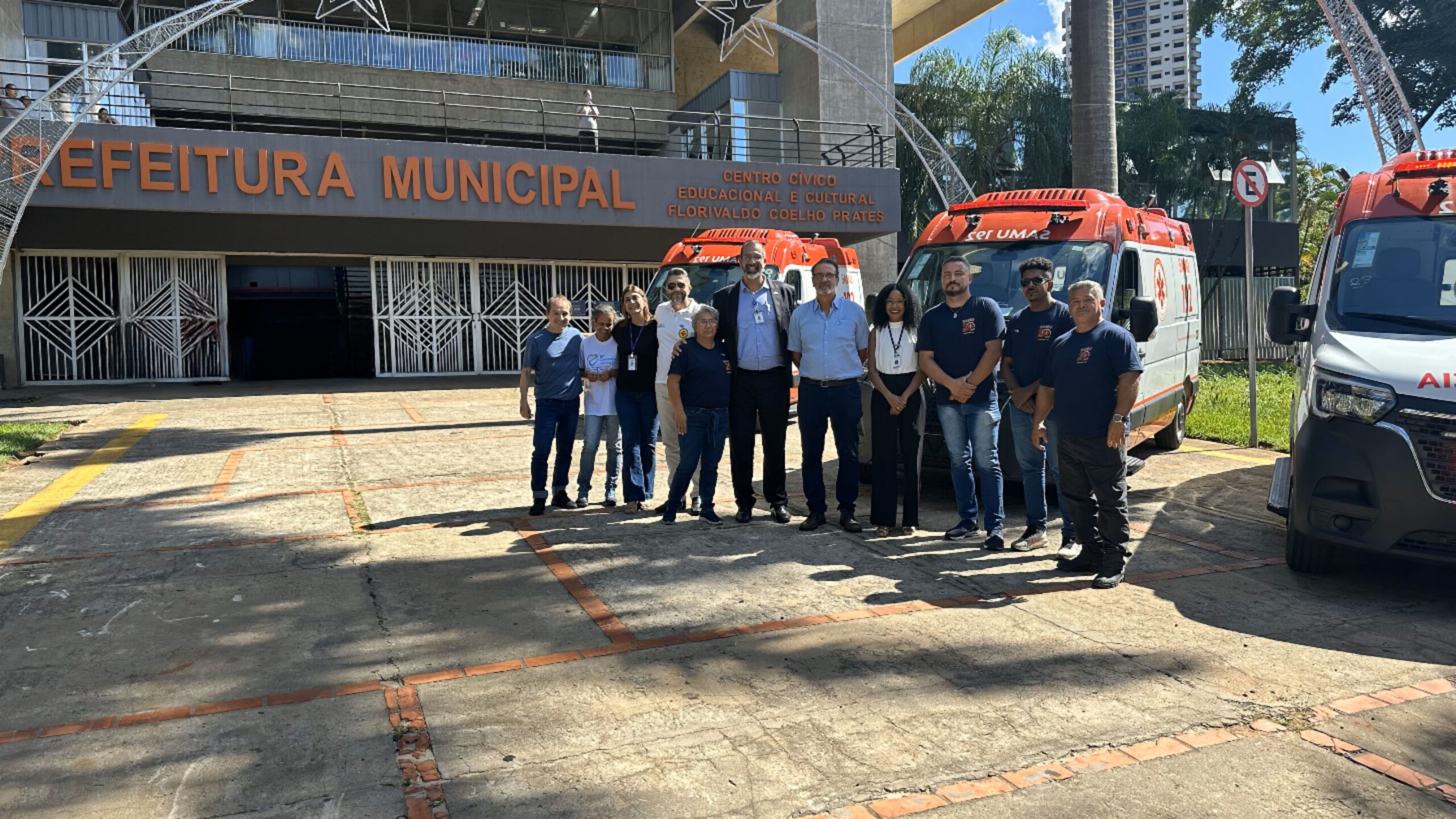 A imagem mostra um grupo de pessoas em pé na frente da Prefeitura Municipal de Florivaldo Coelho Prates. Duas ambulâncias estão estacionadas ao lado do grupo. As pessoas estão sorrindo para a câmera. A maioria está vestindo roupas casuais. Há uma placa na Prefeitura que diz "Centro Cívico Educacional e Cultural Florivaldo Coelho Prates". O local parece ser uma área externa pavimentada, com uma construção de fundo. Há árvores e uma parte de um prédio visível ao fundo. A iluminação sugere que a foto foi tirada durante o dia. Há uma sombra de um poste visível na parte inferior da imagem. O asfalto apresenta algumas marcas e rachaduras.